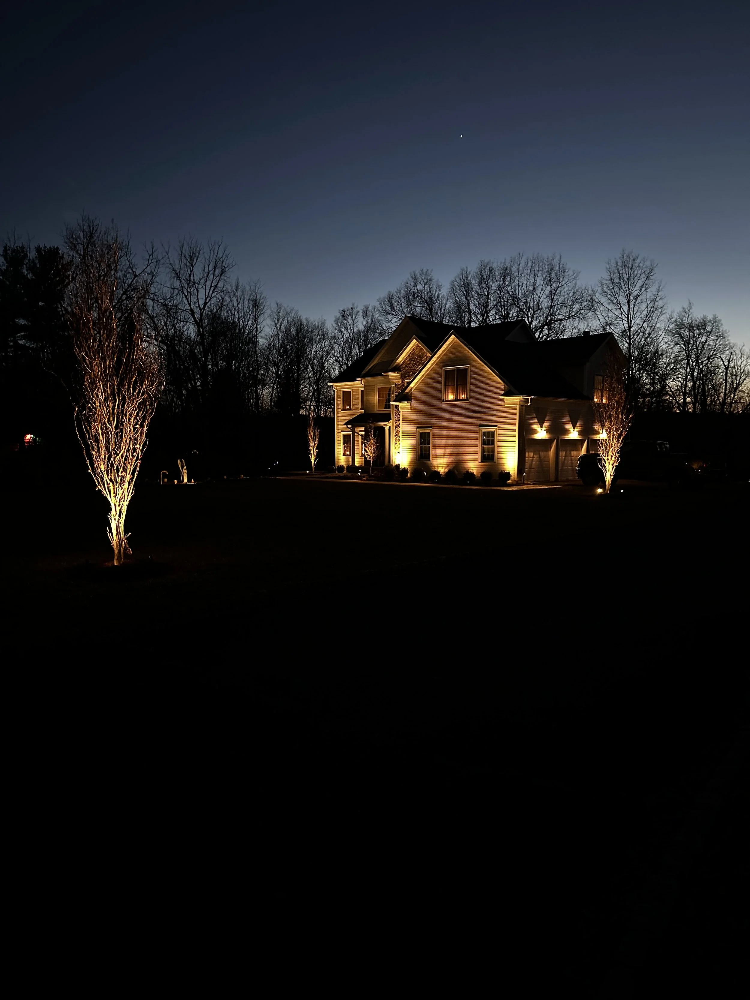 A house at night with exterior lights illuminating the house and surrounding trees, under a dark sky with a visible star.