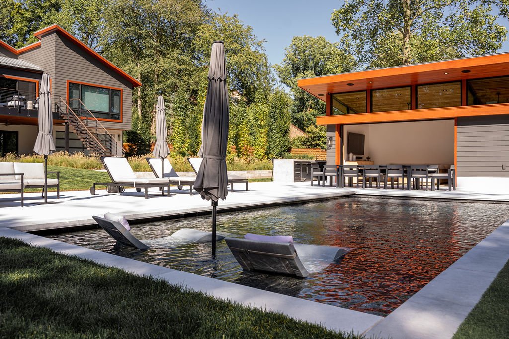 Modern backyard with a pool, chaise lounges, umbrellas, outdoor dining area, and a two-story house with large windows and wooden accents.