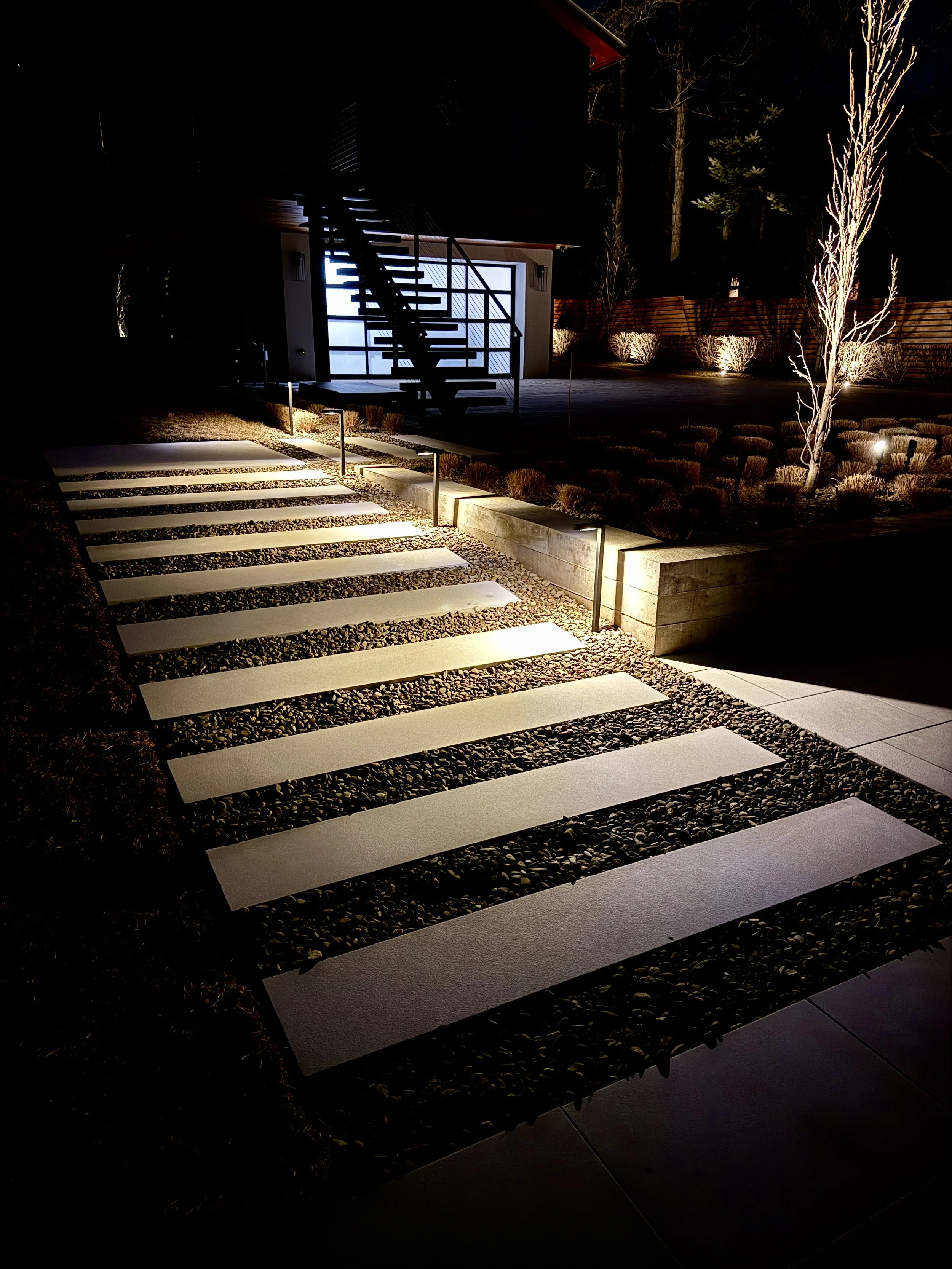 Night view of a modern outdoor walkway with white stepping stones, small ground lights, and a staircase leading to a house with a brightly lit garage, surrounded by landscaped yard with trees and shrubs.