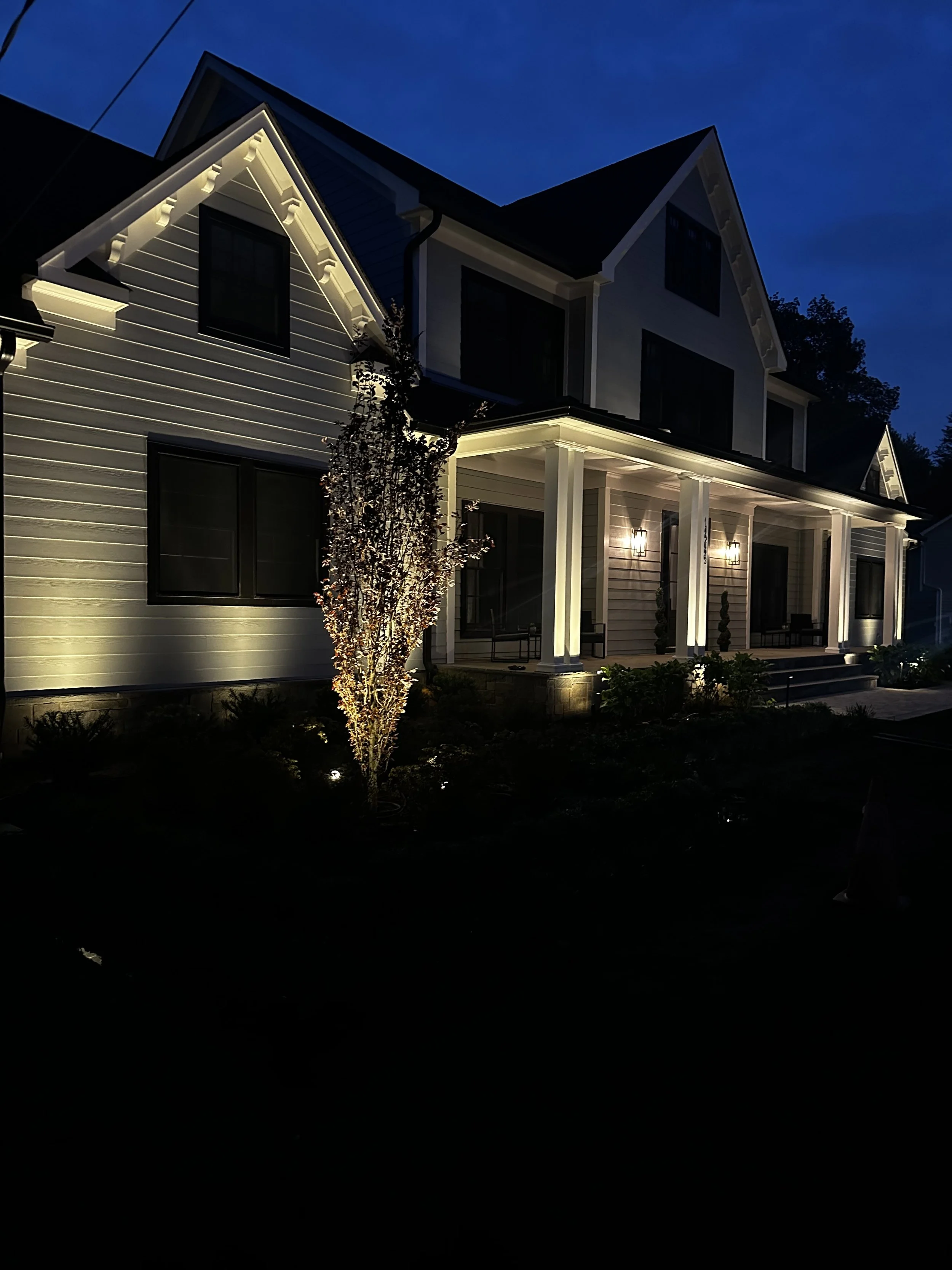 A large, modern house illuminated with exterior lighting at night, featuring a porch with columns, a small tree in front, and a dark sky in the background.