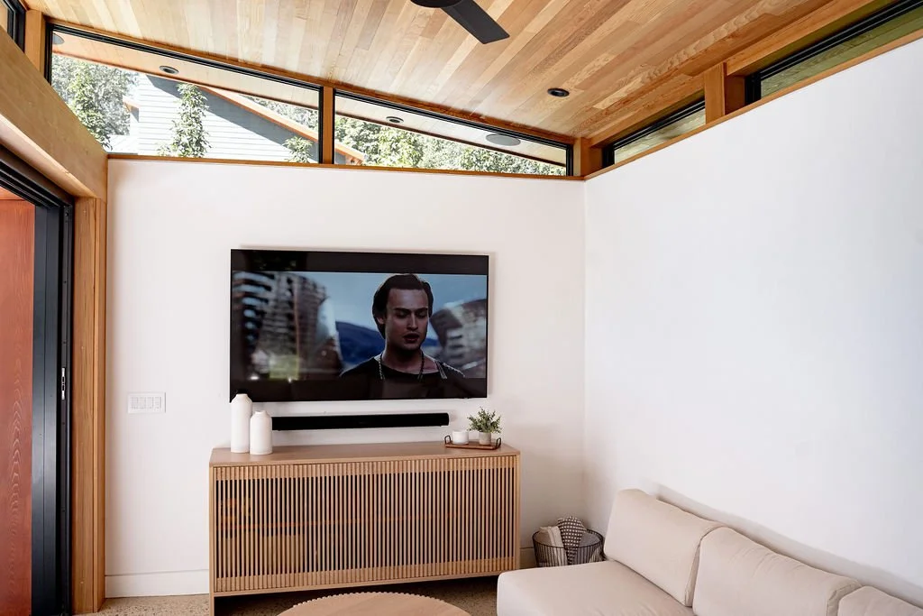 A modern living room with a flat-screen TV on a wooden cabinet, a beige sofa, and windows with natural light and greenery outside.
