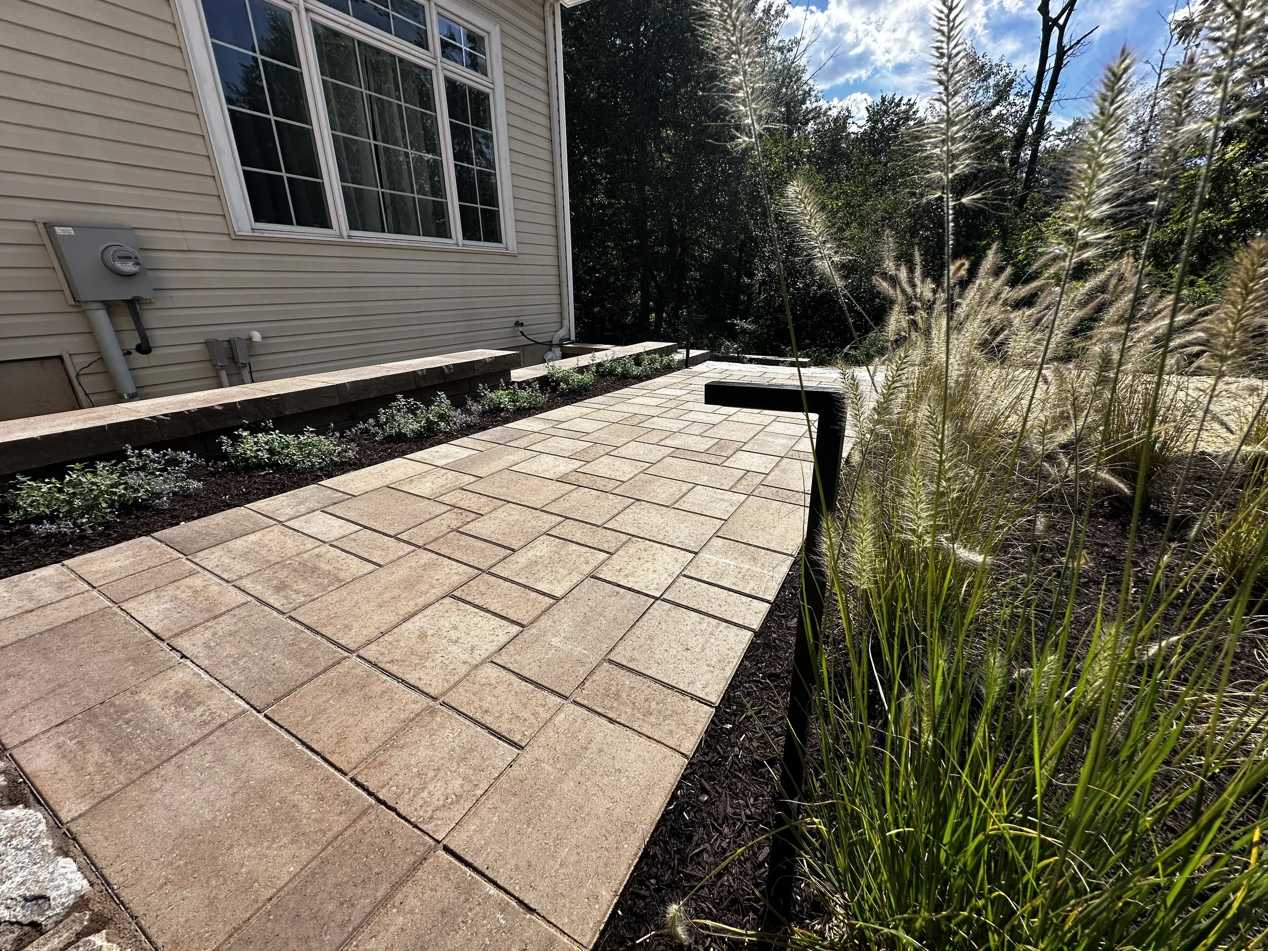 A newly paved brick pathway leading away from a house with beige siding, surrounded by landscaping and ornamental grasses, under a partly cloudy sky.