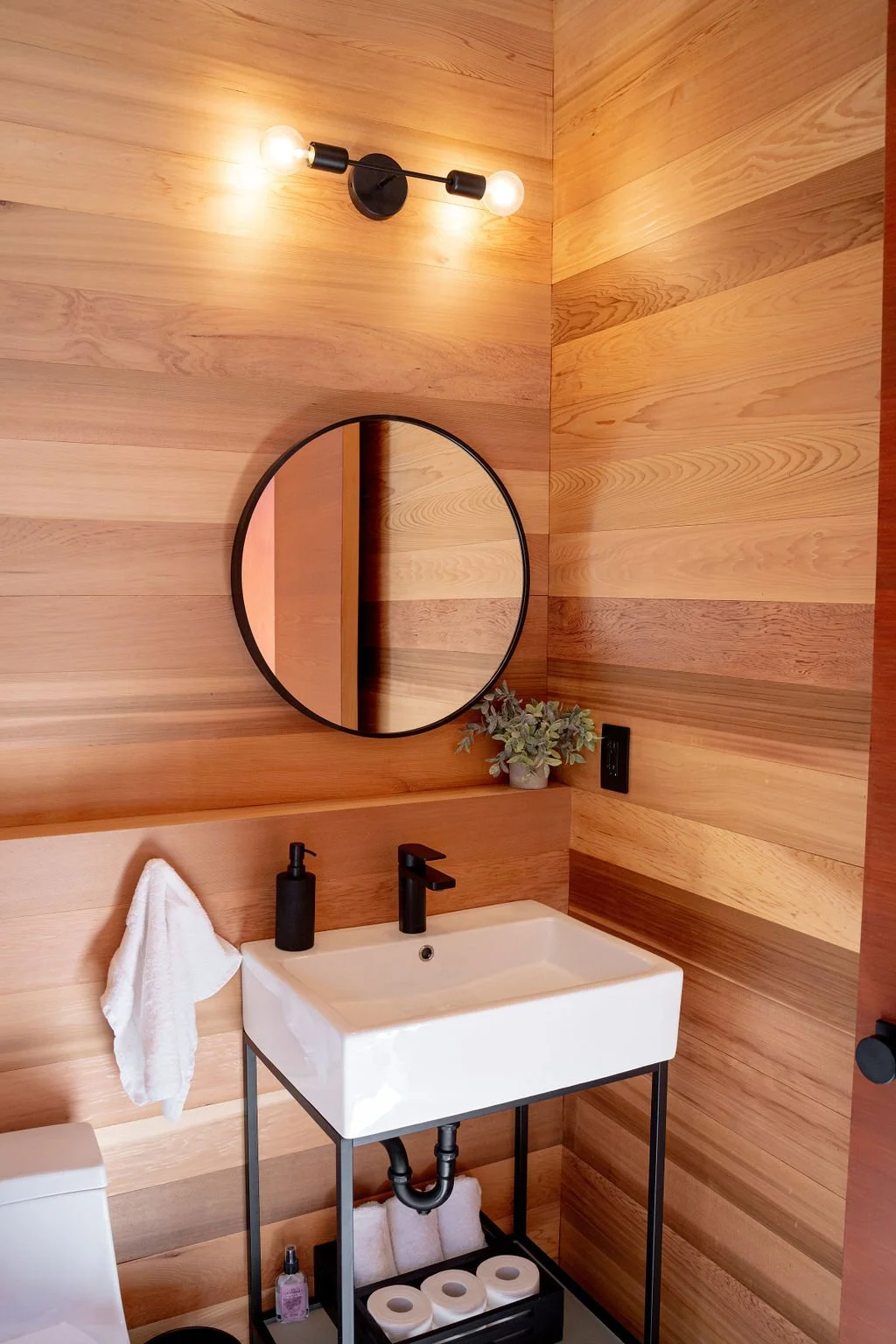 A modern bathroom with wood-paneled walls, a round mirror, black light fixture, small plant, and white sink with black fixtures.