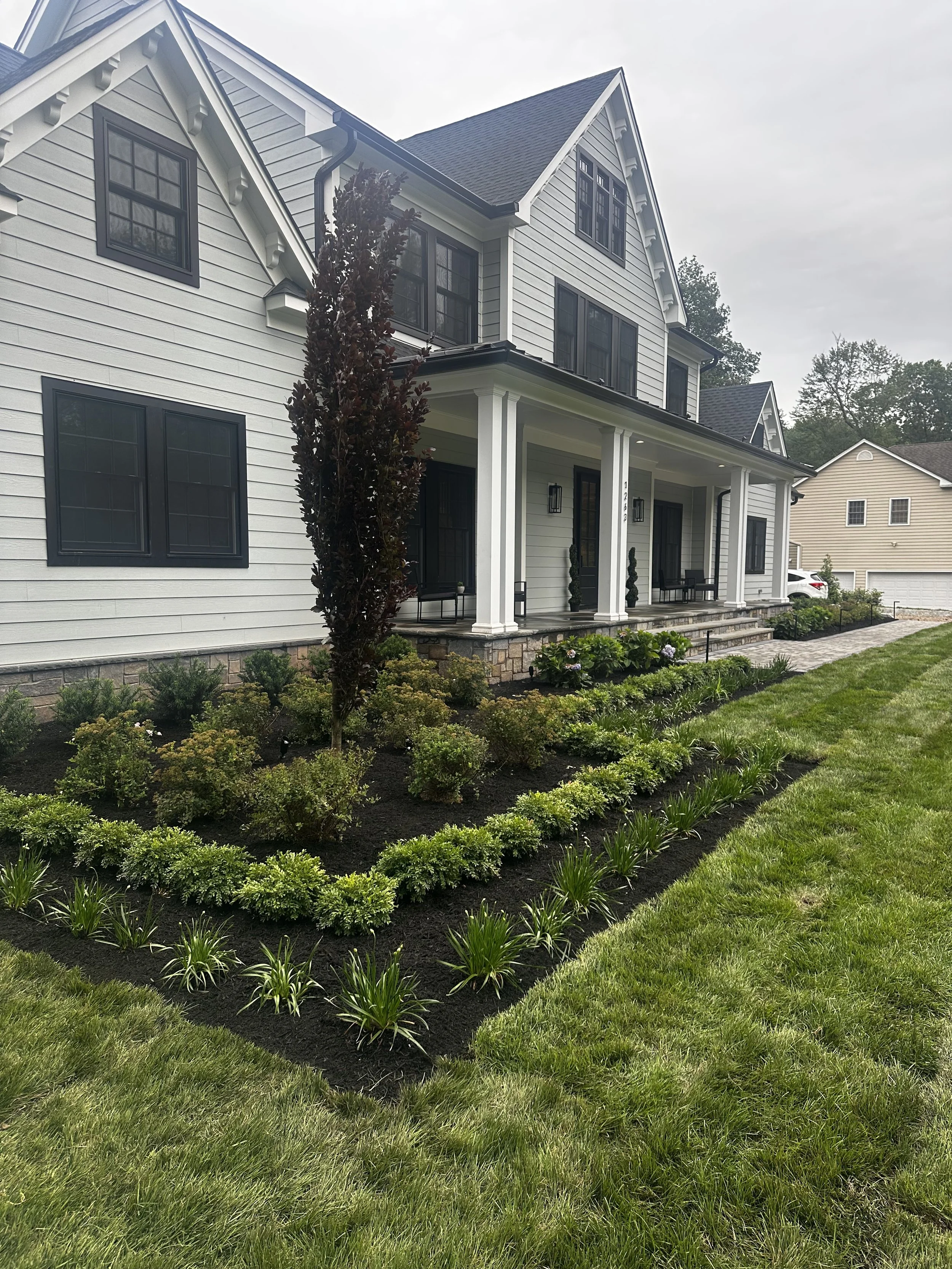 Front yard landscaping with manicured lawn, shrubs, flower beds, and a tall tree in front of a white house with black window frames and a covered porch.