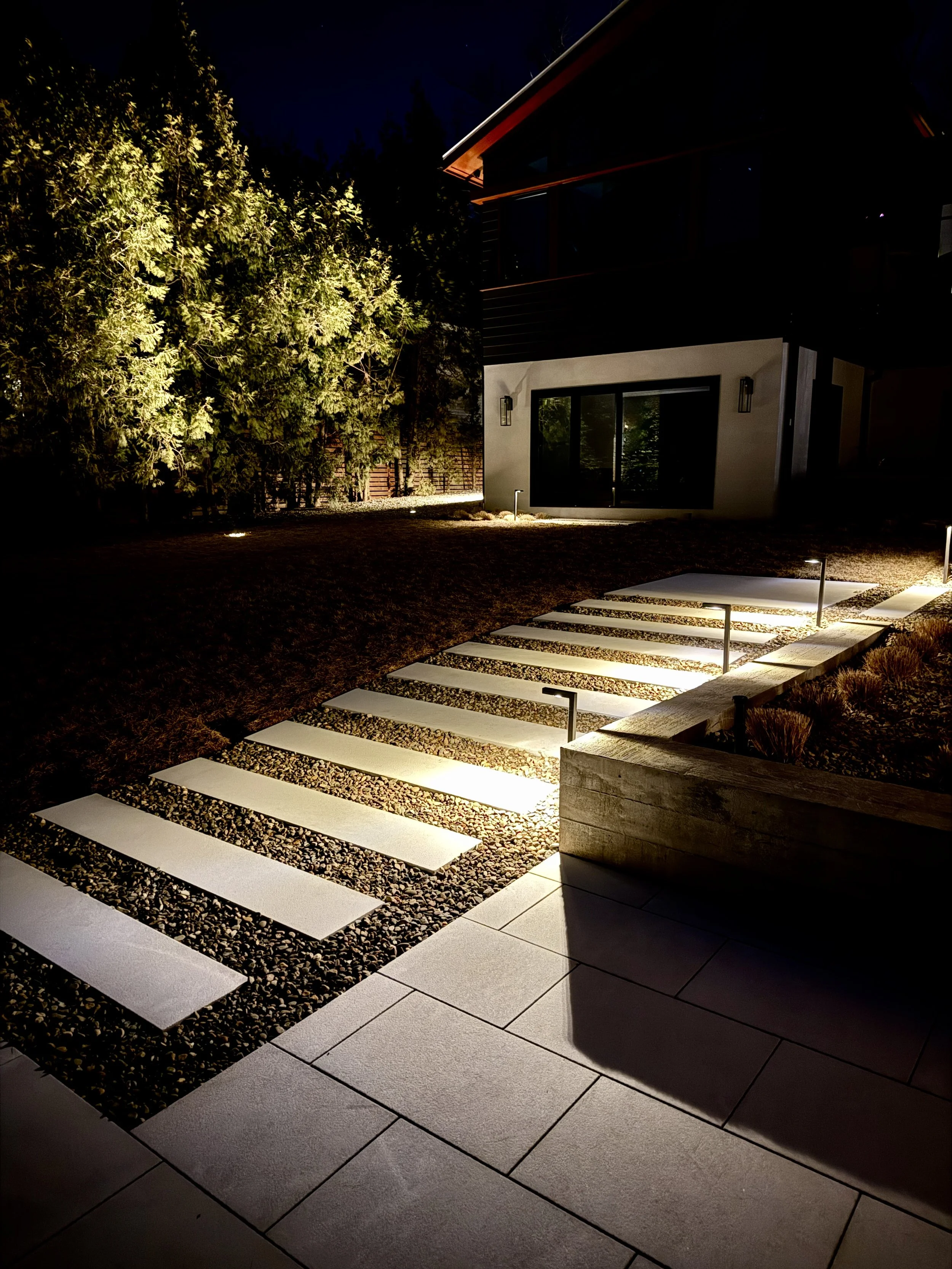 Nighttime exterior view of a modern house with a front yard, illuminated pathway with white rectangular stepping stones, trees, and a balcony.