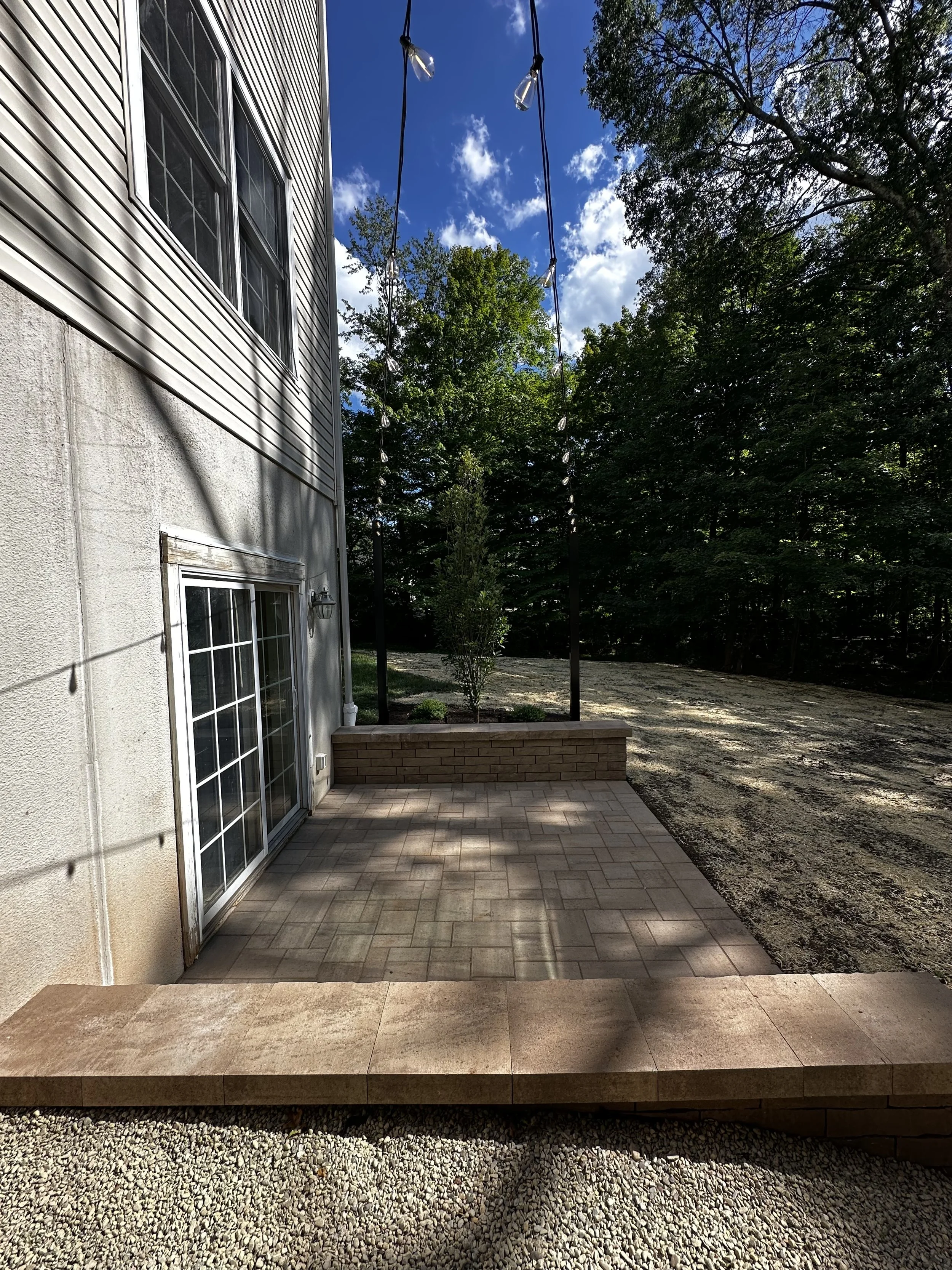 Newly constructed back patio with brick pavers, a small raised brick border, string lights hanging above, and a backyard with trees and dirt ground.