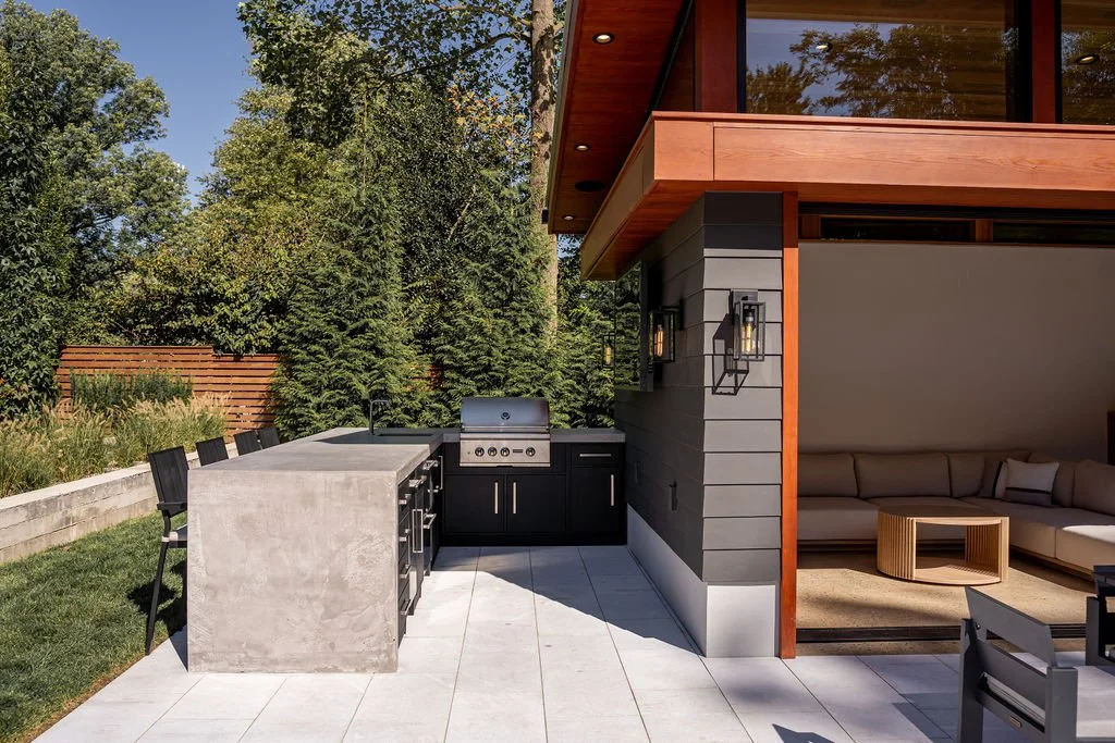 Outdoor kitchen and patio with built-in grill, concrete countertop, black cabinets, and a seating area with a sectional sofa under a wooden-beamed roof, surrounded by trees.
