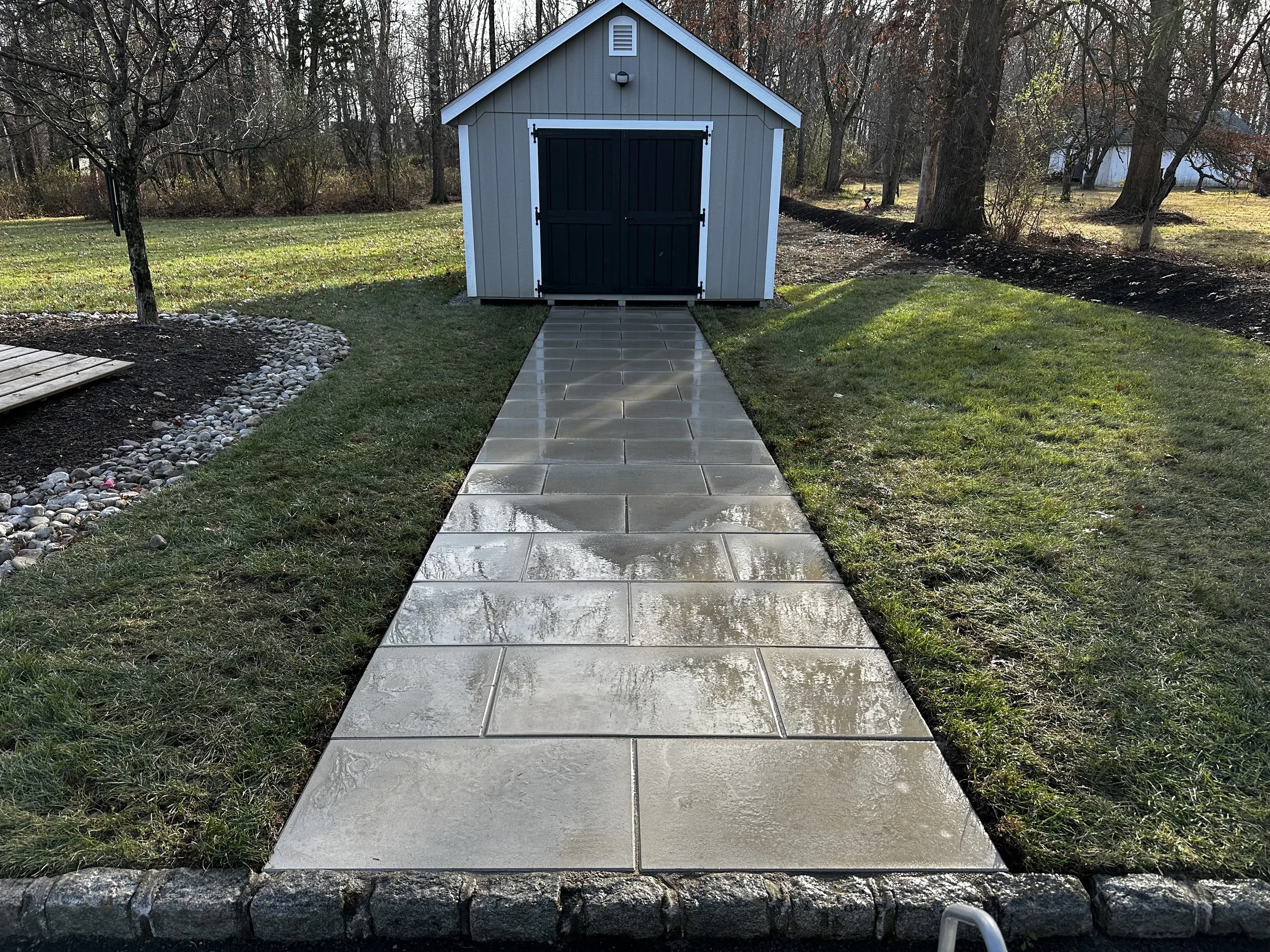 A concrete pathway leading to a small gray shed with black doors in a backyard, with grass on both sides and trees in the background.