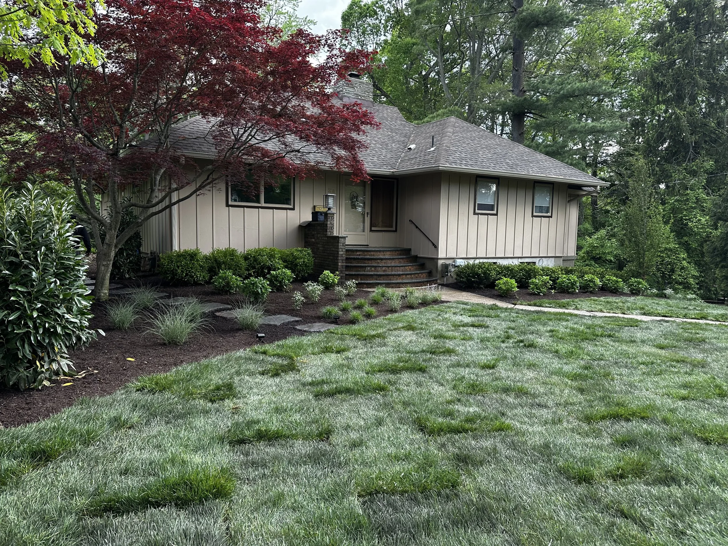 front view of a beige single-story house with a gray shingle roof, surrounded by green trees and landscaped garden with bushes, grass, and a small walkway.