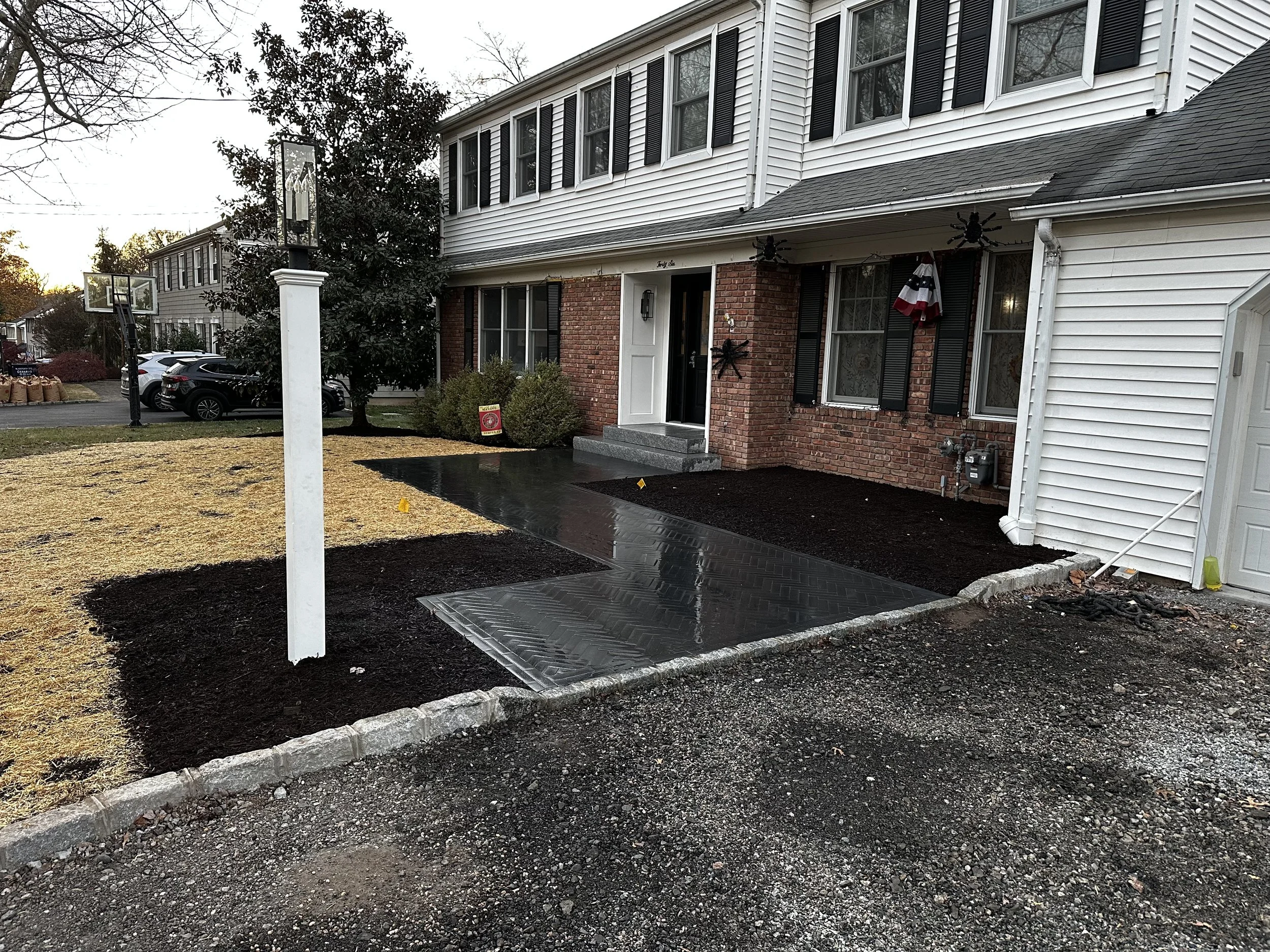 Front yard of a house under renovation with fresh black asphalt walkways and a small patch of mud beside a brick and white siding house, with a white mailbox, decorative spider, and American flag decoration.
