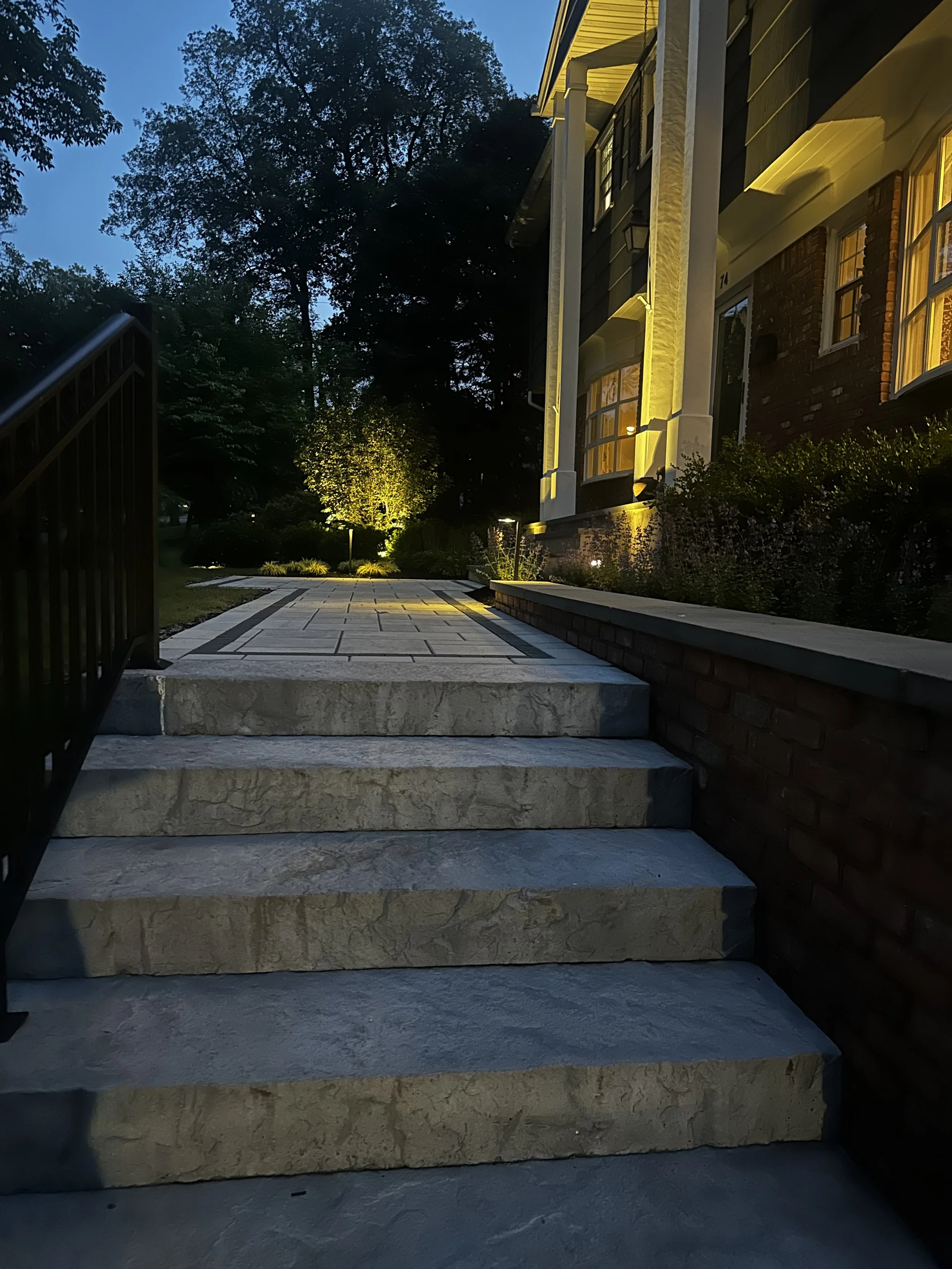 Stone steps leading to a house entrance at dusk, with exterior lighting on the house and trees illuminated in the background.