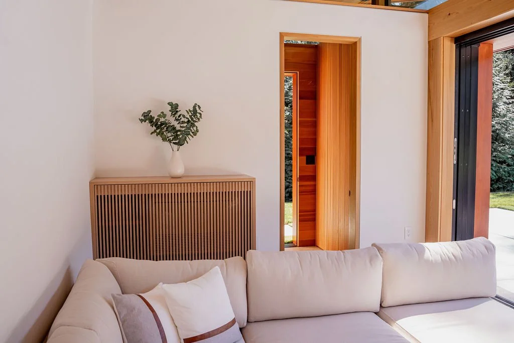 Modern living room with white sofa, wooden vent cover, white vase with green foliage, and large window/door opening to outdoor greenery.