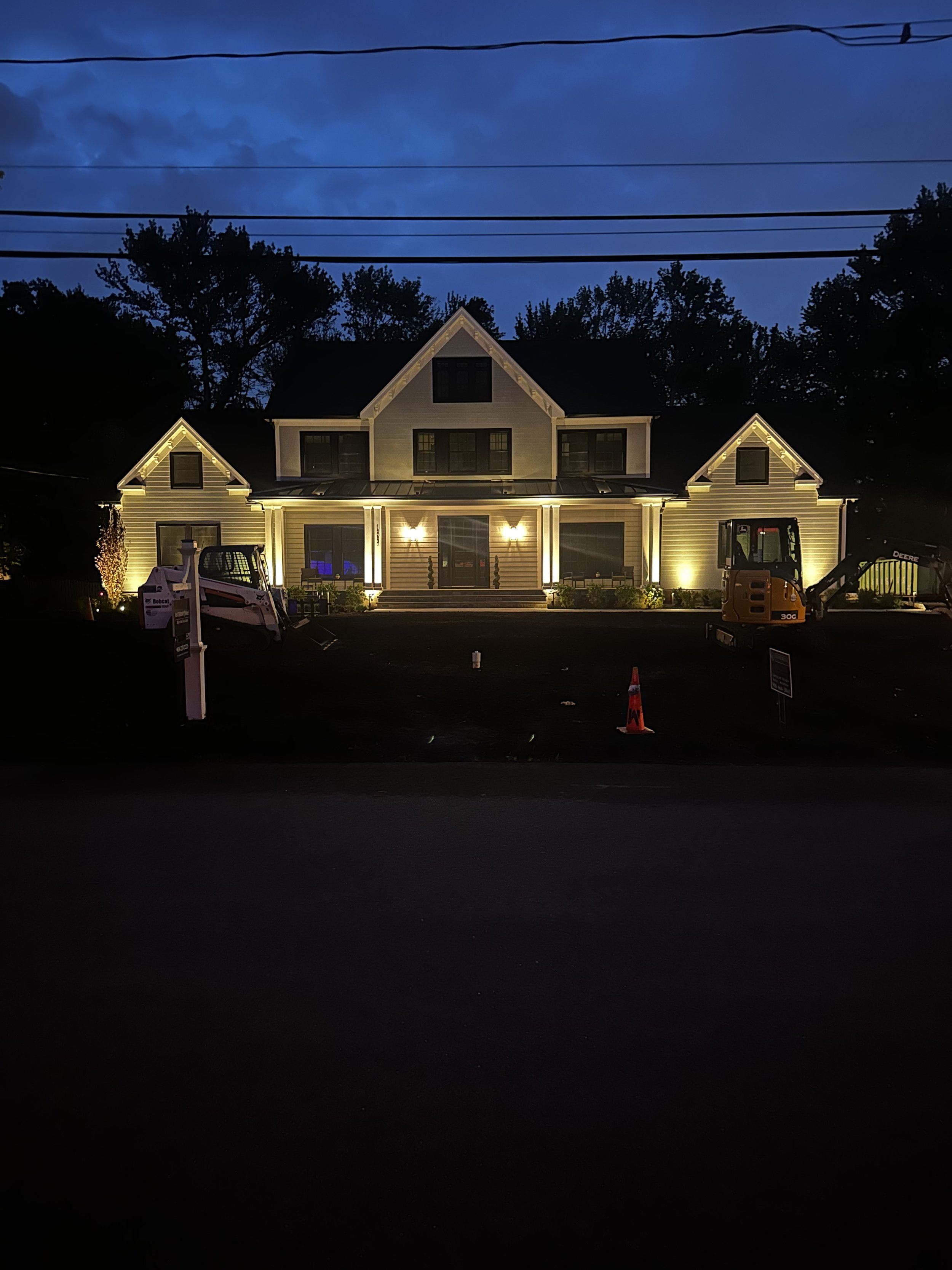 Illuminated house with construction equipment in front during dusk, trees and power lines in the background.