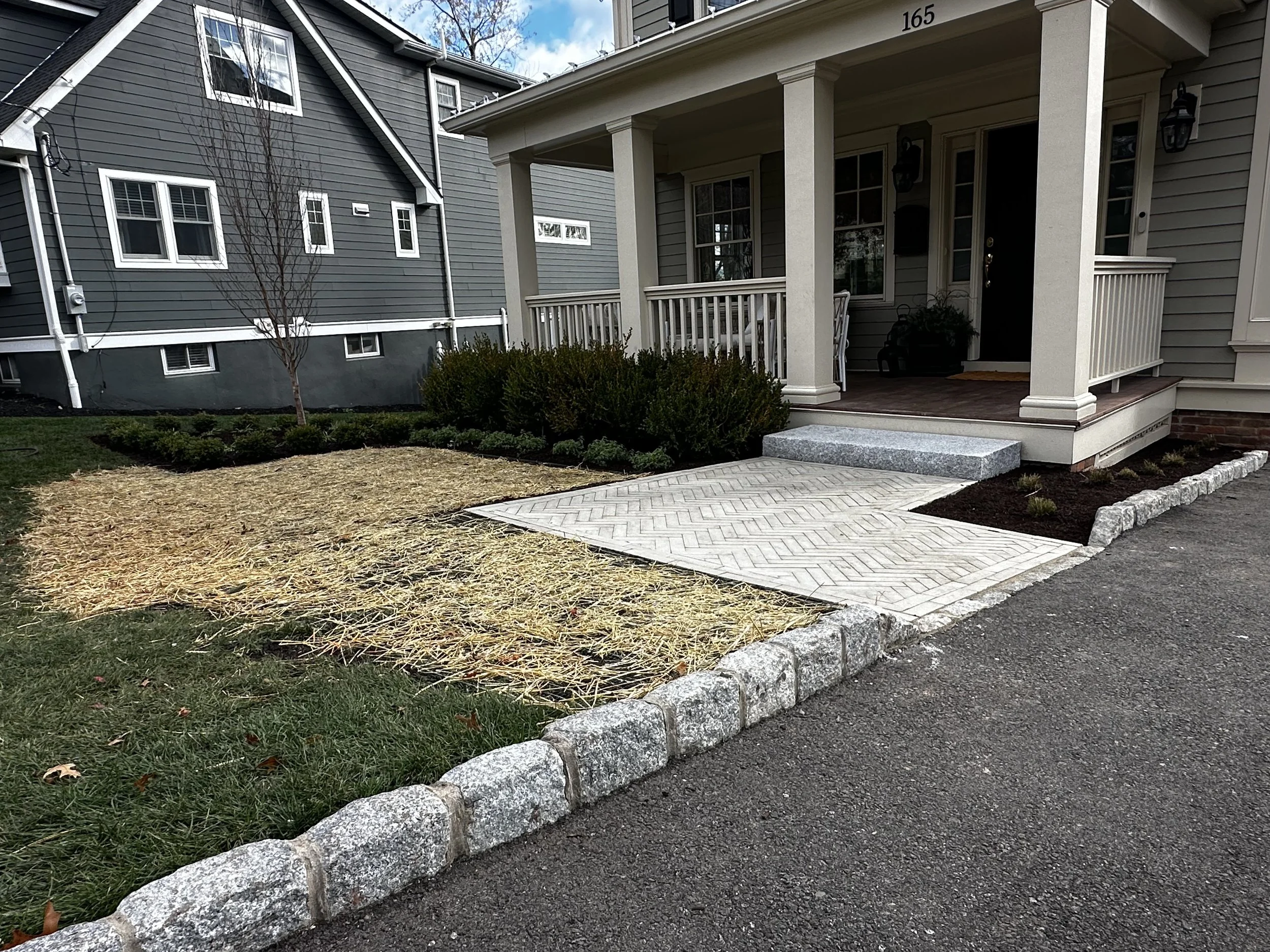 Front porch of a house with a small set of stairs, a paved walkway, and landscaping including bushes and a small, leafless tree, adjacent to a driveway with a stone border.