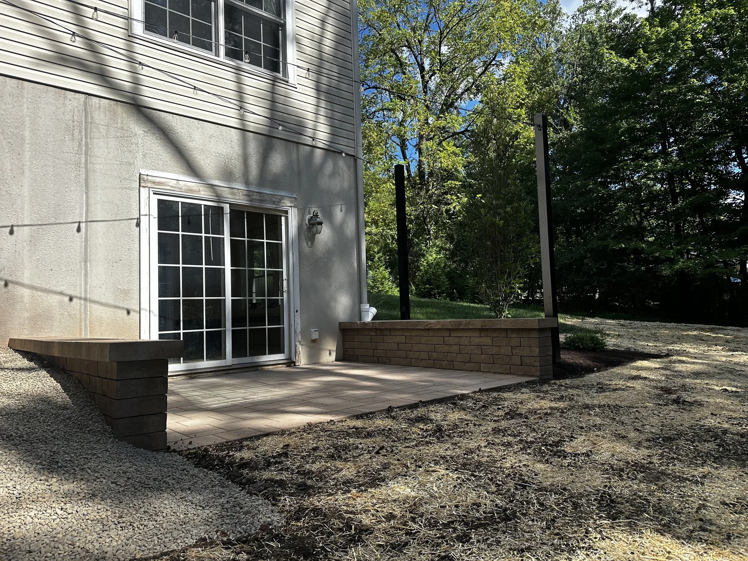 Backyard patio area with a sliding glass door leading into a house, surrounded by trees and greenery, with a portion of the patio bordered by brown bricks and a gravel section