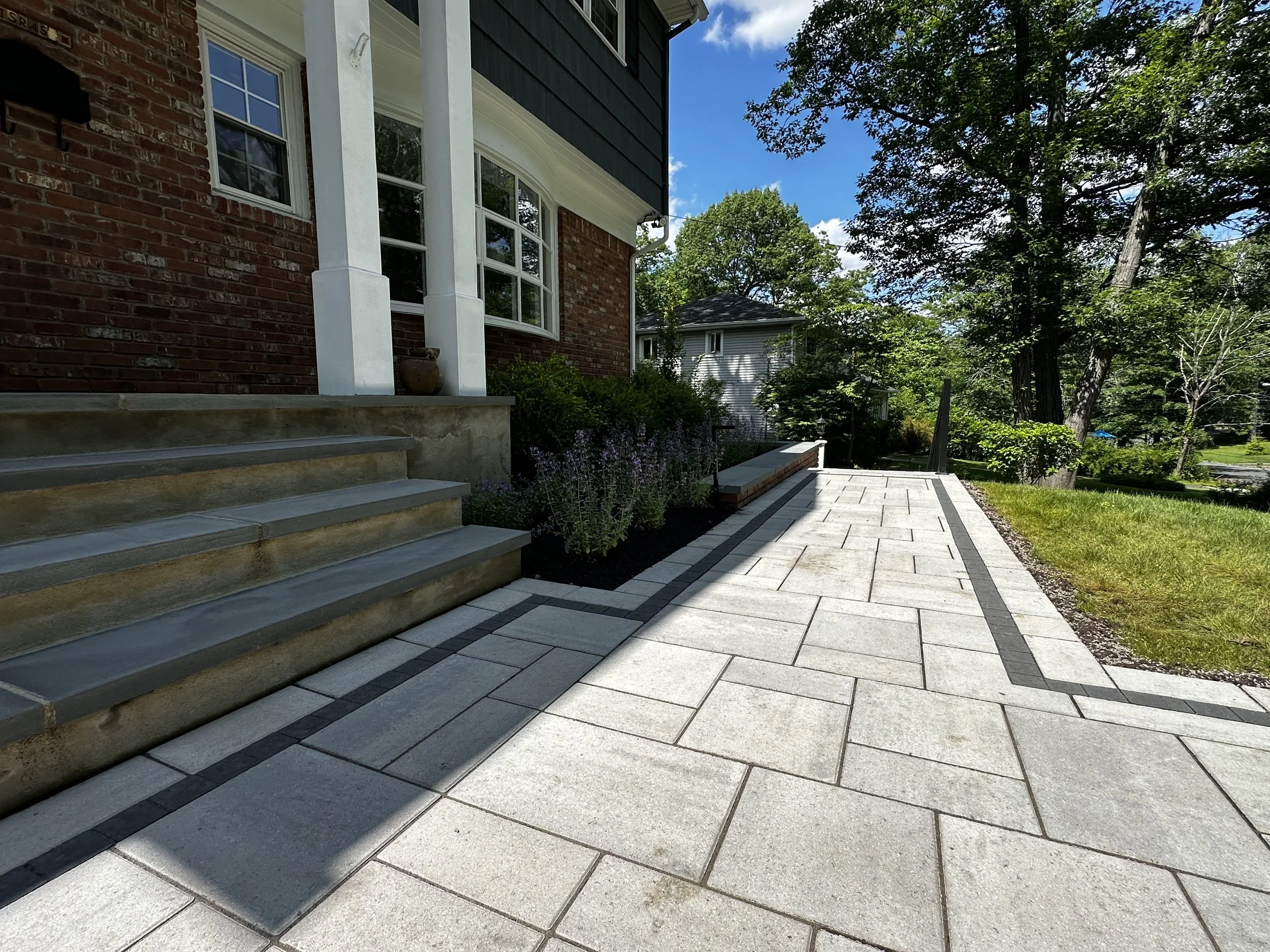 A front yard with a brick house, concrete steps, a paved walkway, landscaping, and trees on a sunny day.
