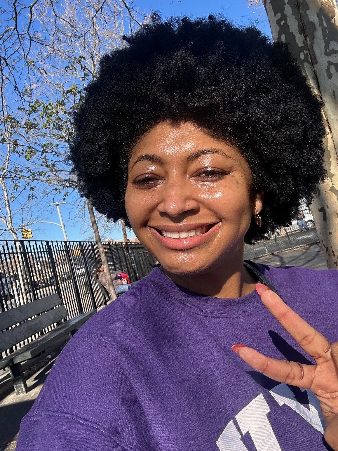 Young woman in a purple sweatshirt, standing in a park in New York City, smiling.
