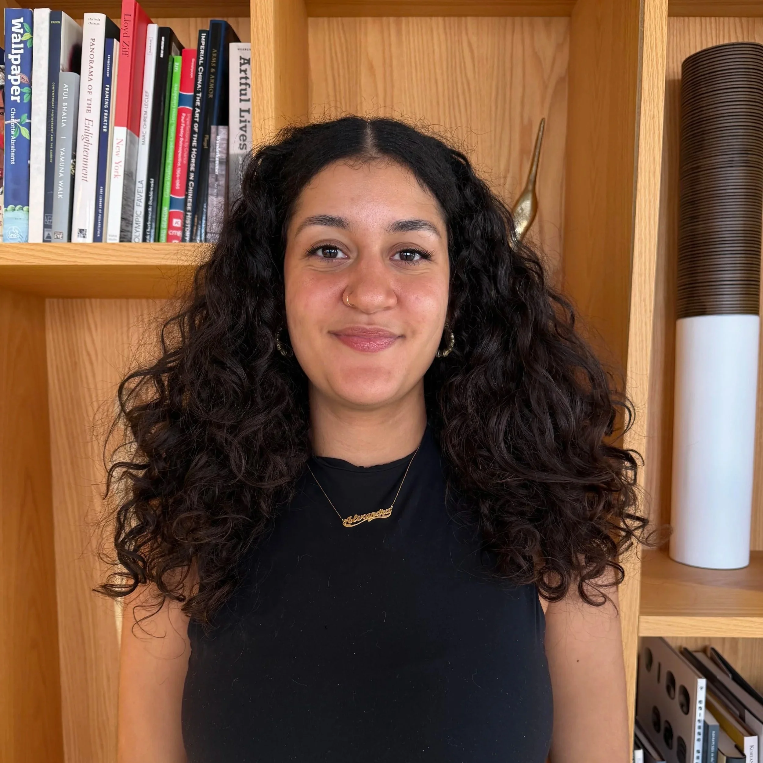 young woman with logn dark curly hair in black tshirt in front of a bookcase