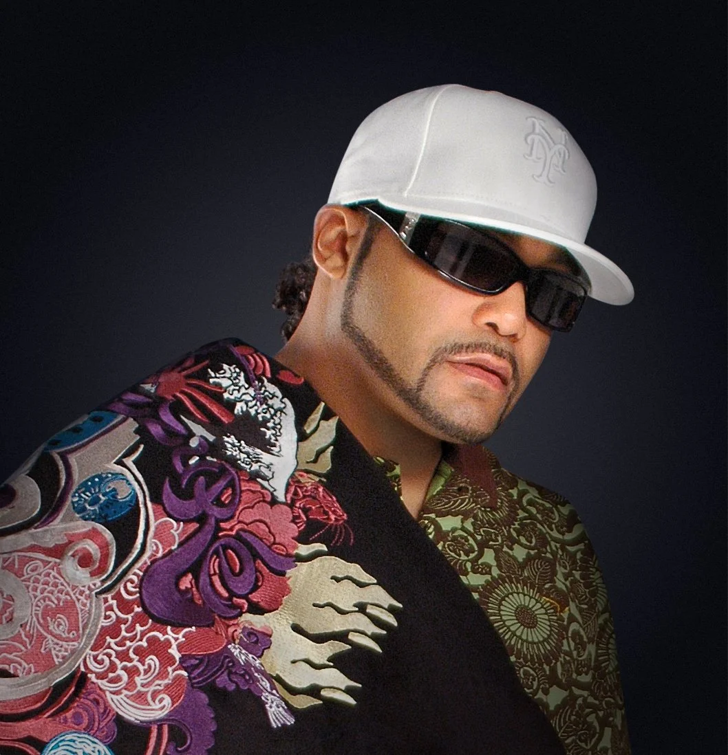 A man wearing a white baseball cap, black sunglasses, and a colorful patterned shirt against a dark background.