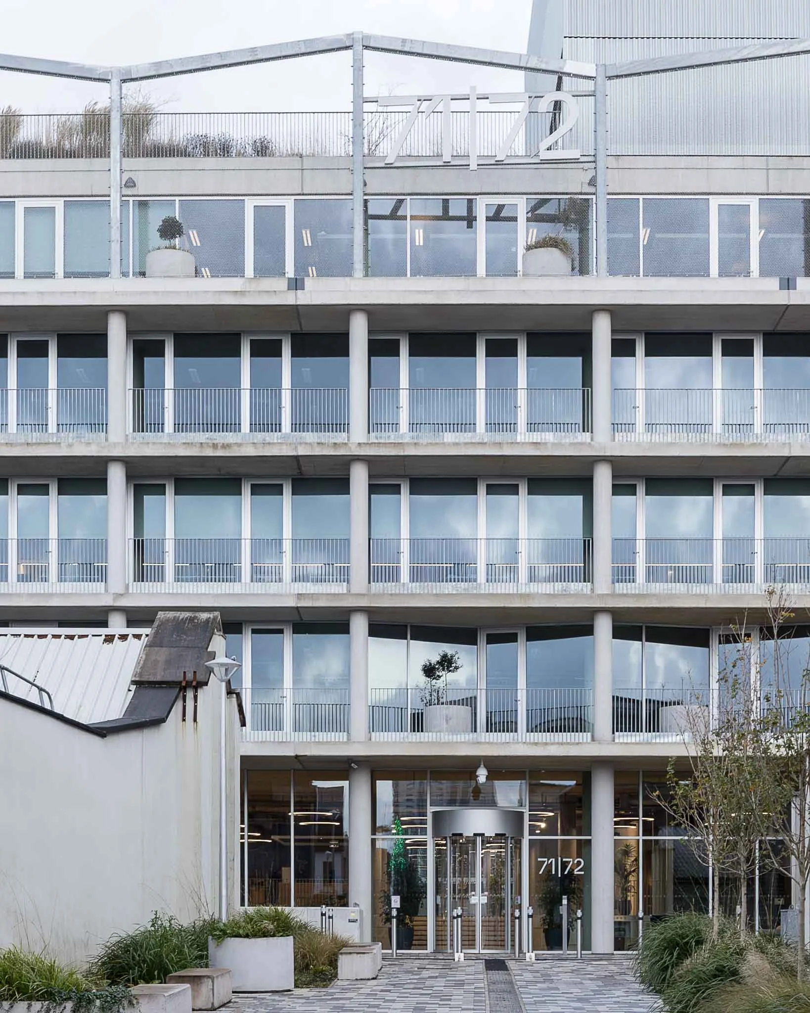 Modern multi-story office building with large glass windows and balconies, decorated for Christmas with a small Christmas tree inside visible through the front door, and a festive wreath on the entrance door. Exterior landscaping features plants and 