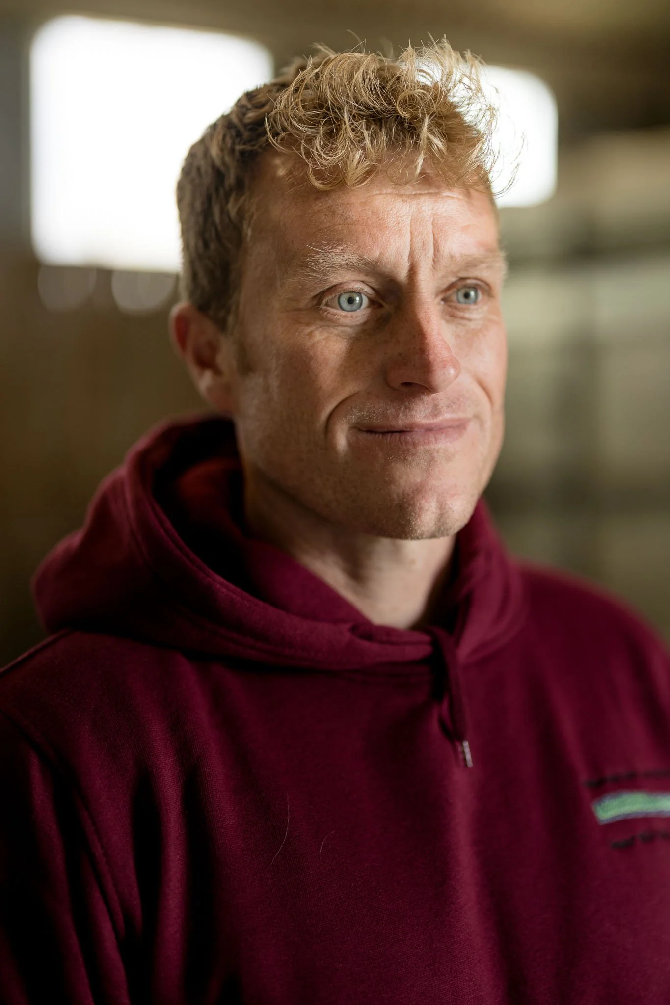 A man with curly blonde hair and blue eyes wearing a maroon hoodie, looking to the side with a slight smile.