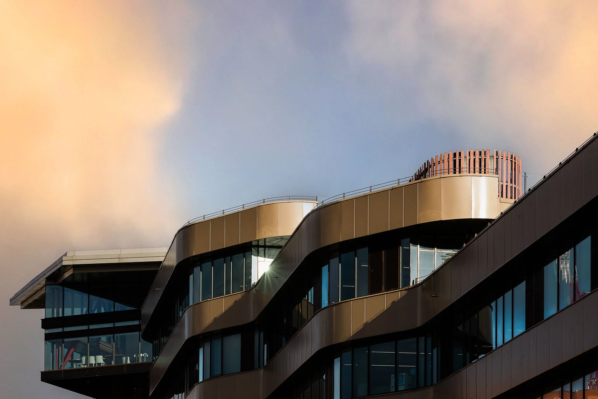 Modern multi-story building with large glass windows and rounded balconies, illuminated by sunlight against a cloudy sky.