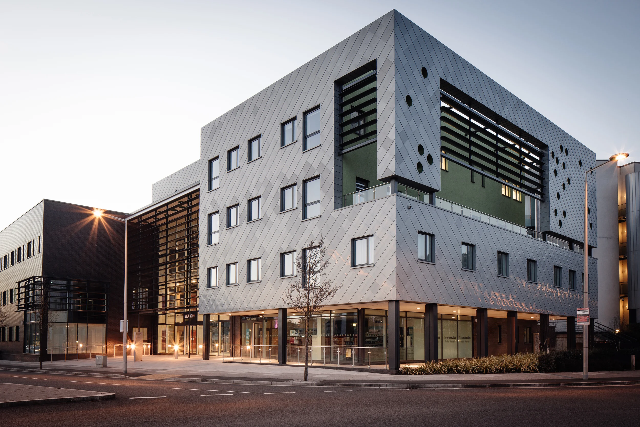 Modern multi-story building with a sleek gray and green exterior, large windows, and contemporary architecture. Streetlights and a tree are visible outside at dusk.