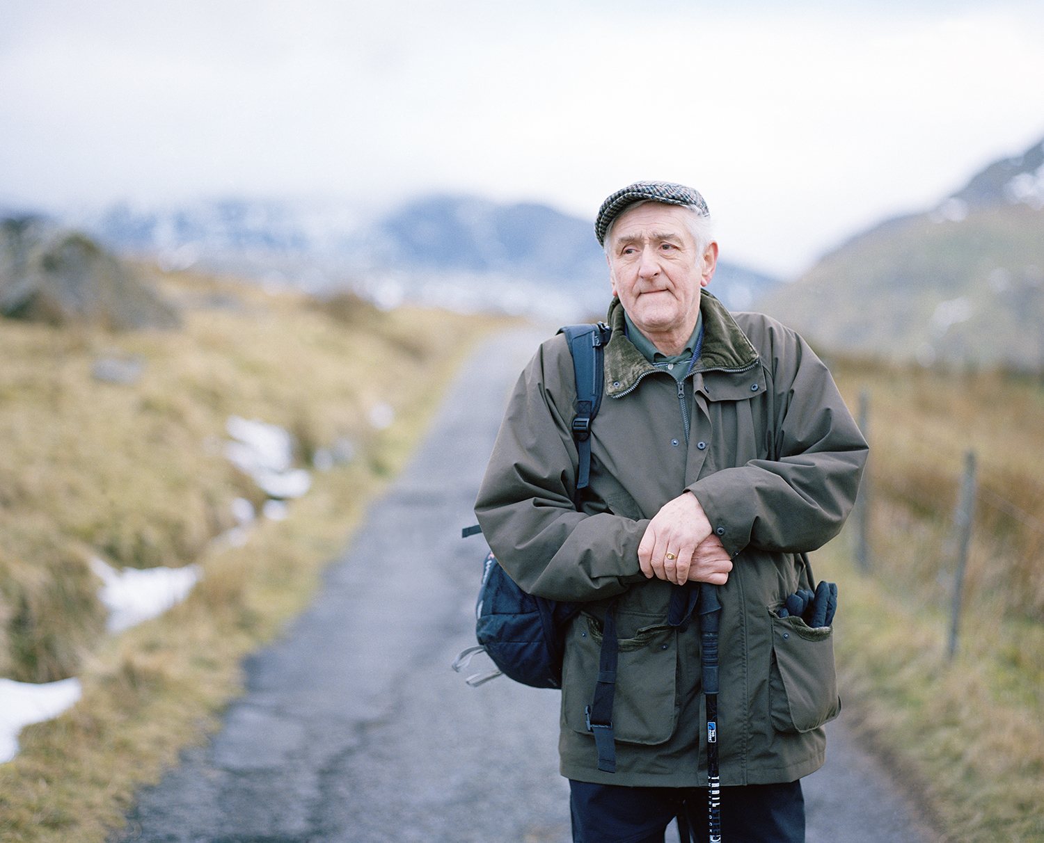 An older man with gray hair wearing a flat cap, green jacket, and carrying a backpack, stands on a narrow path in a mountainous landscape with cloudy sky and snow patches in the background.