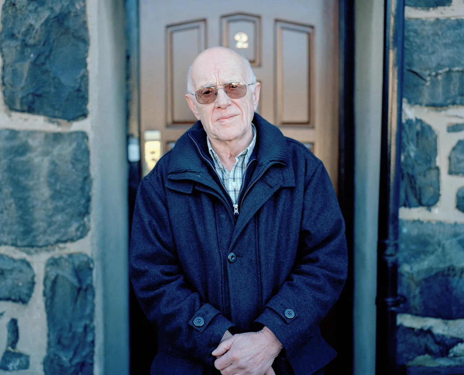 An elderly man with glasses and a blue coat standing in front of a wooden front door framed by stone walls.