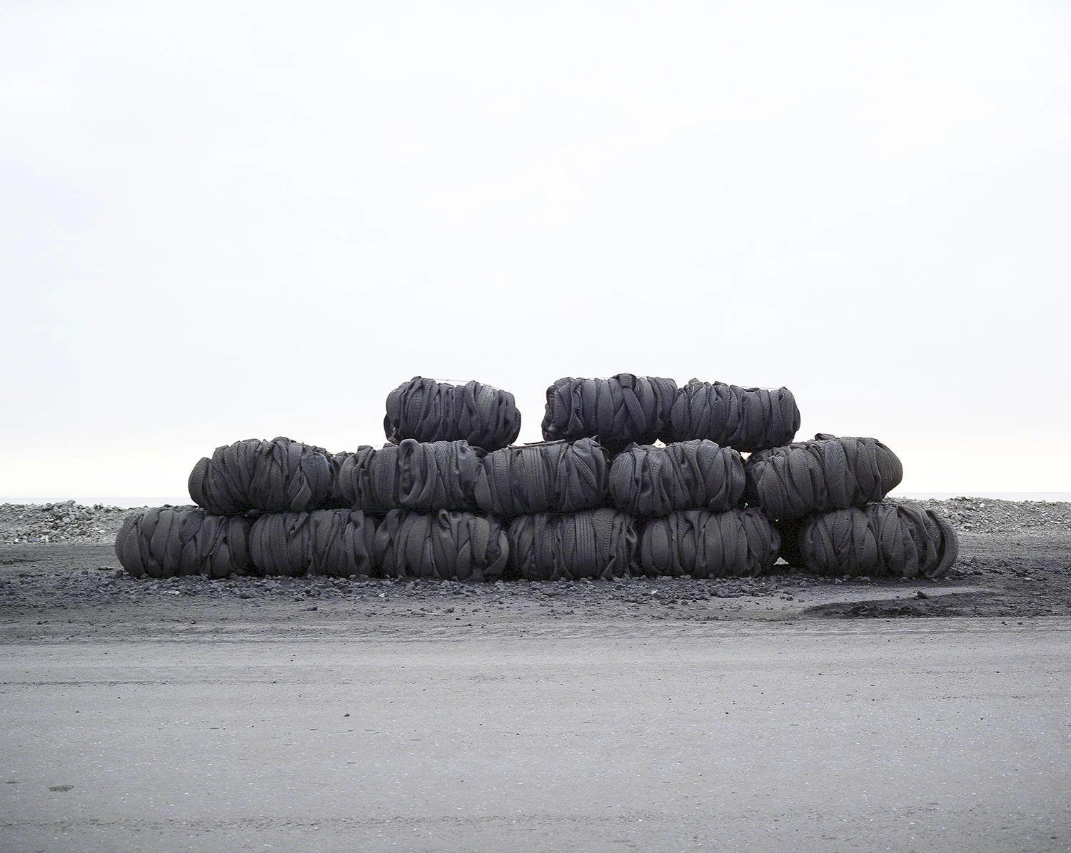 Stack of large, black rubber tires on gravel in a barren, open landscape with a cloudy sky.