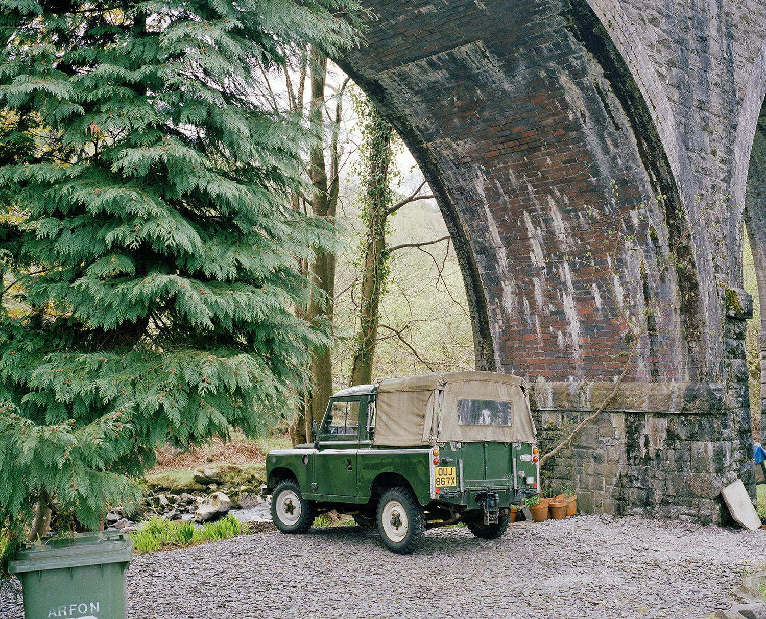 A small vintage green vehicle with a beige canvas top parked under a large brick arch bridge in a natural setting with trees and a stream nearby.