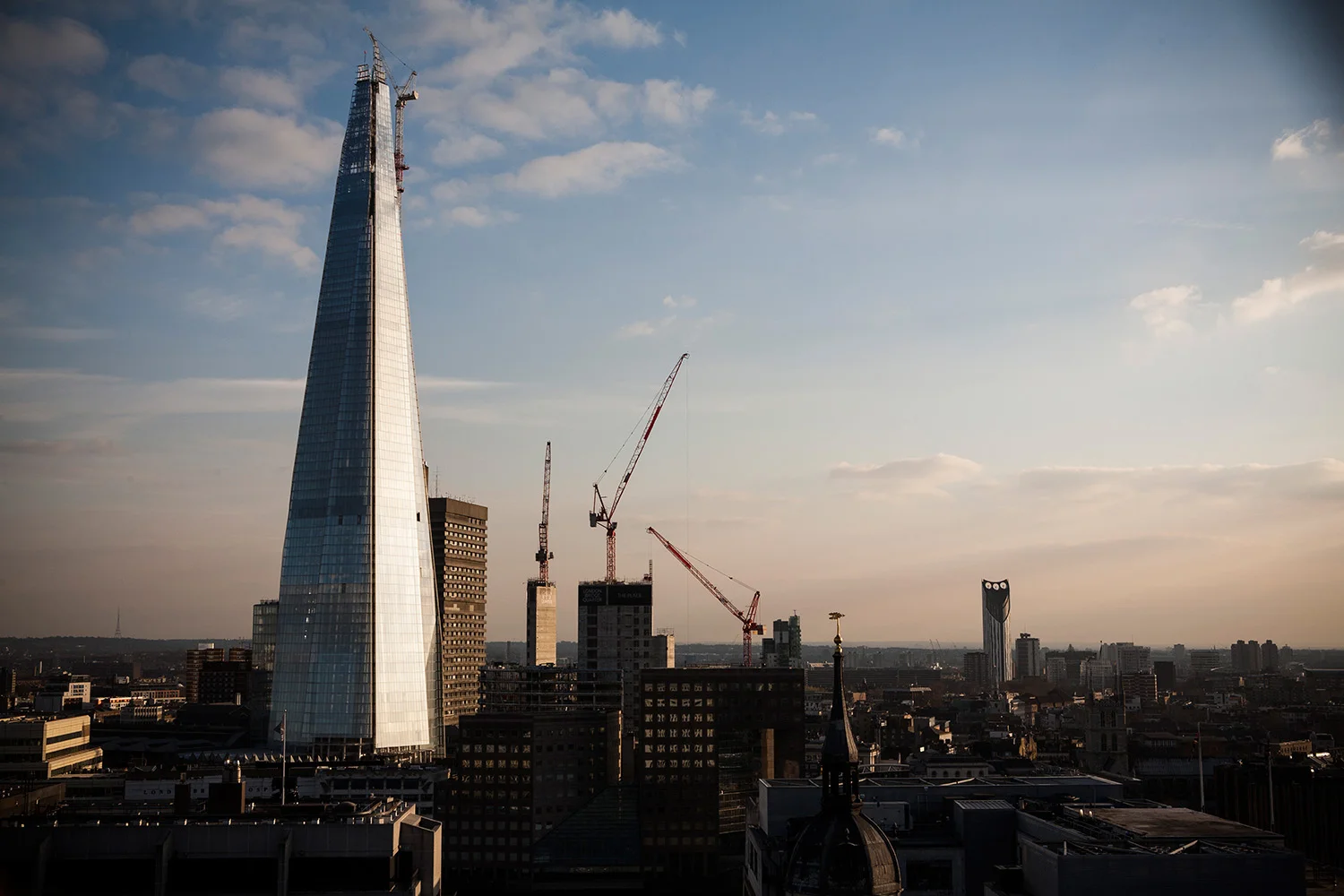 City skyline featuring The Shard skyscraper, under construction cranes, and other buildings, with a partly cloudy sky at sunset.