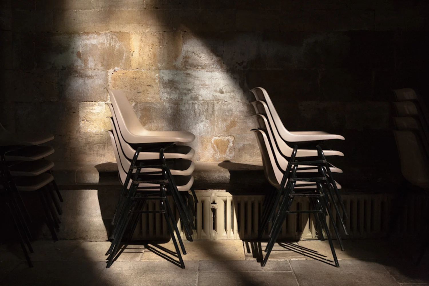 Stacked beige chairs on top of black chairs against a stone wall, lit by a spotlight in a dim room.