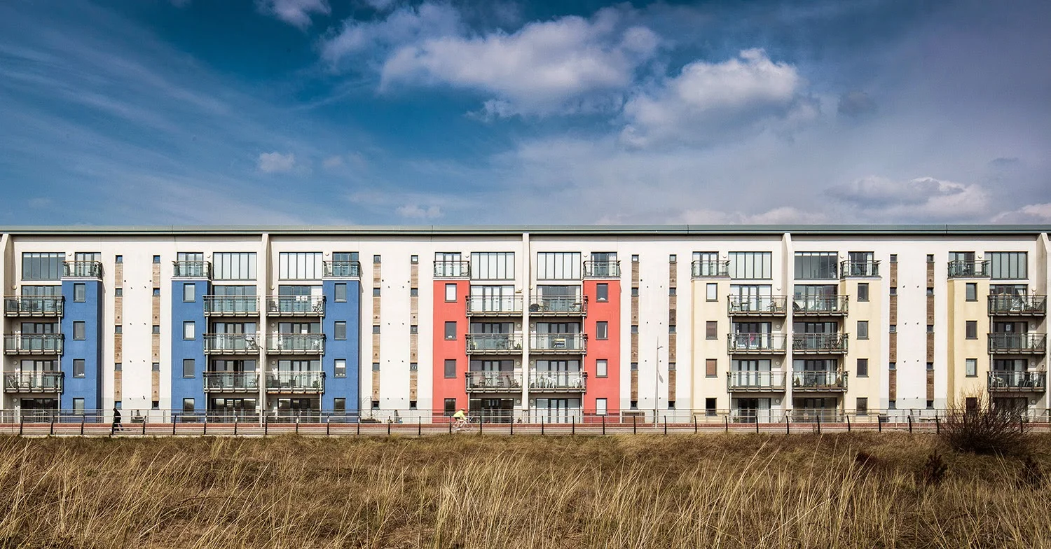 Colorful apartment building with blue, red, and white sections, balconies, and large windows, set against a partly cloudy sky and grassy foreground.