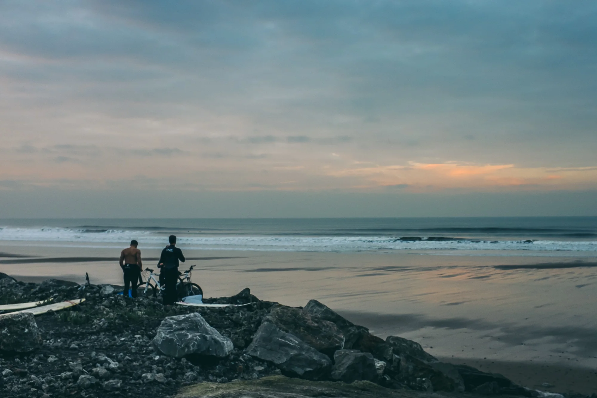Two men standing on a rocky beach, one shirtless and the other wearing a wetsuit, beside their bicycles, looking towards the ocean at sunset.