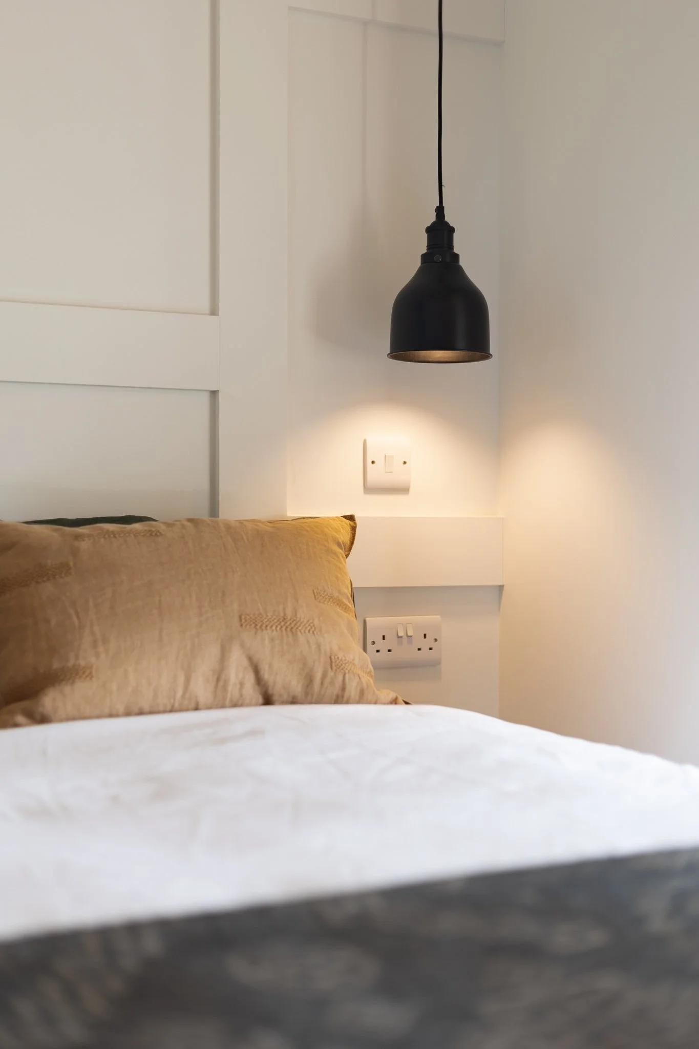 Close-up of a bedroom with a brown pillow, a white bedsheet, a wall-mounted black pendant light, and electrical outlets.