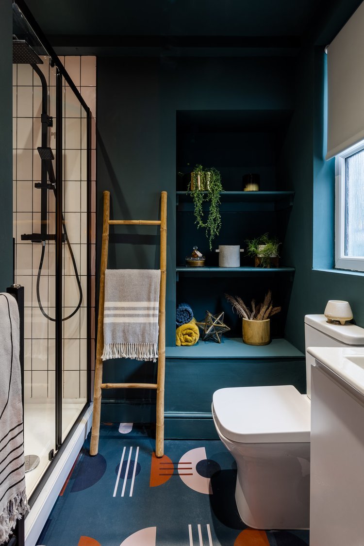 Modern bathroom with teal walls, a walk-in shower with black fixtures and beige tiles, a wooden ladder towel rack with a striped towel, built-in teal shelves holding plants and decorative items, a window with a white blind, a white toilet, and a colorful geometric patterned rug.