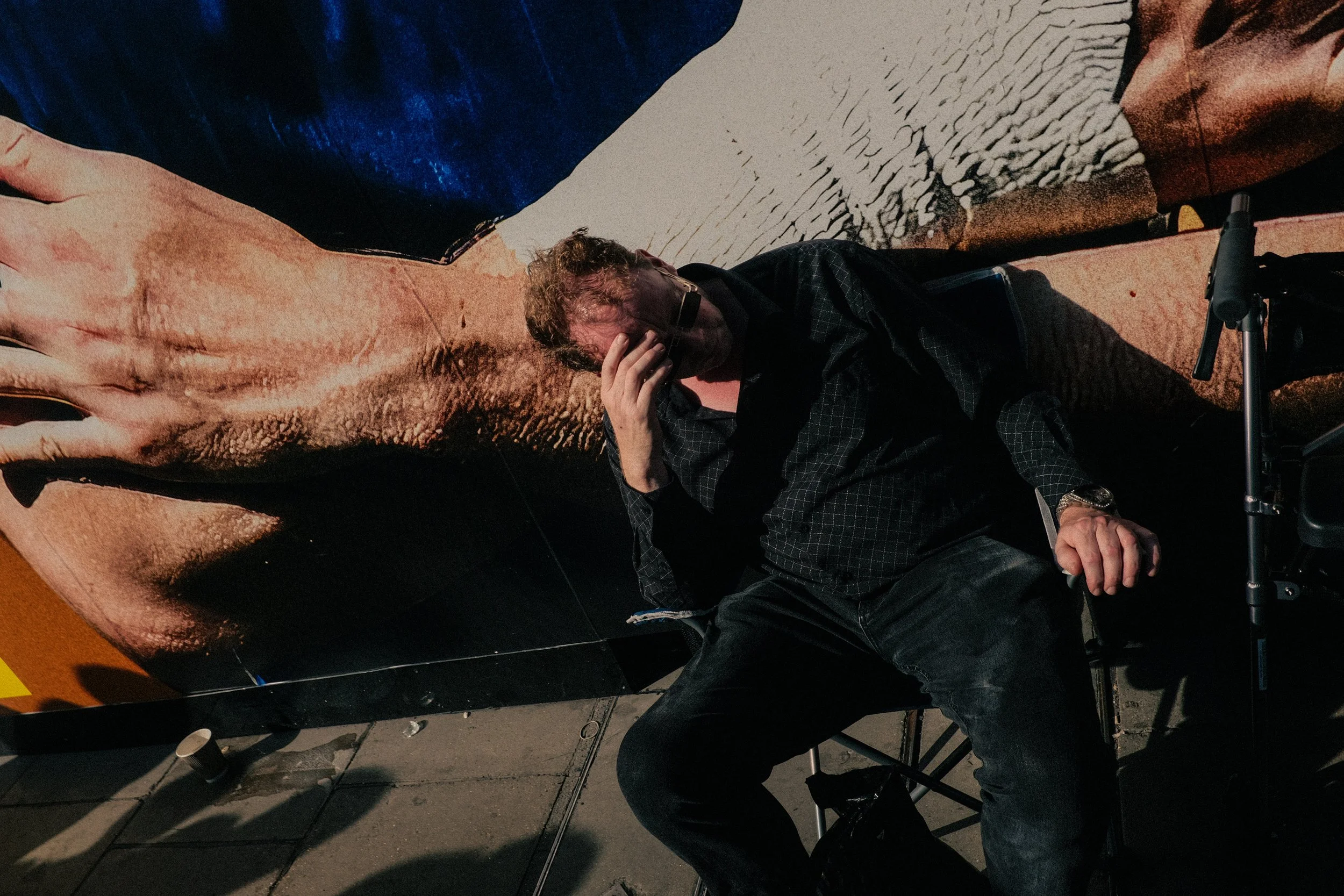 Man sitting on floor reading or resting, surrounded by painted or printed artwork, with a camera tripod nearby.