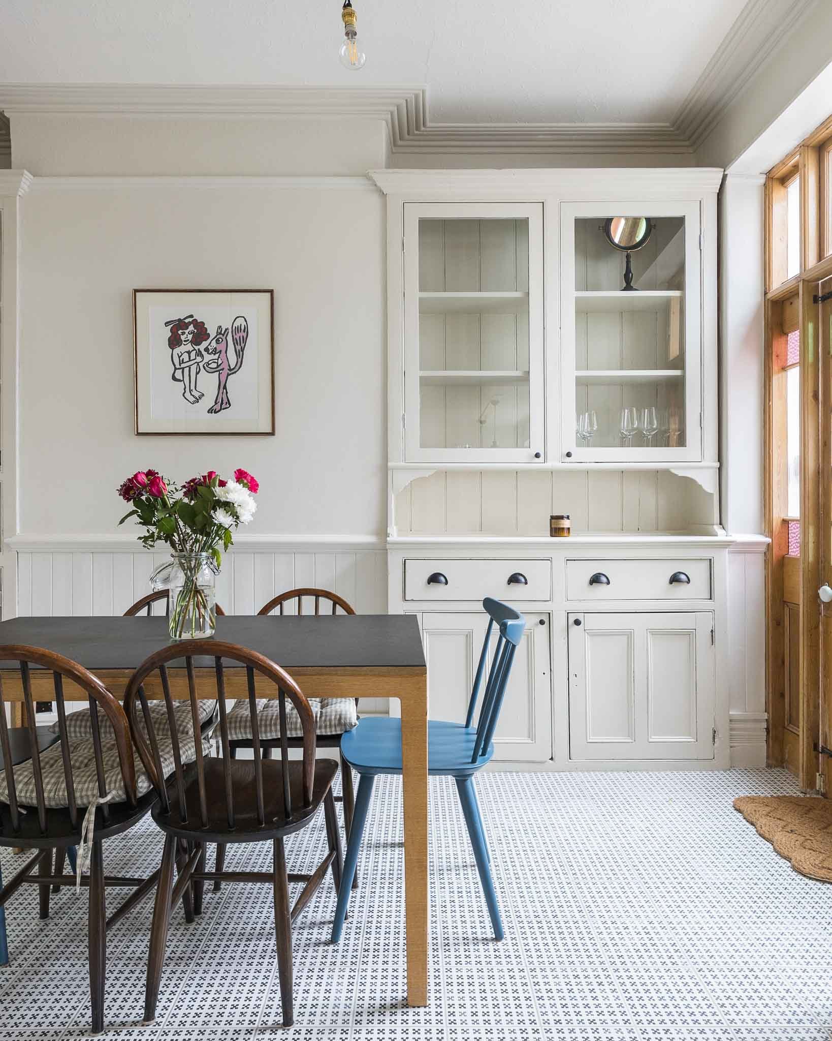 A dining area with a black table and wooden chairs, a blue chair, and a white cabinet with glass doors. A small mirror and a candle are on top of the cabinet, and a framed art piece hangs on the wall. A vase with pink and white flowers is on the tabl