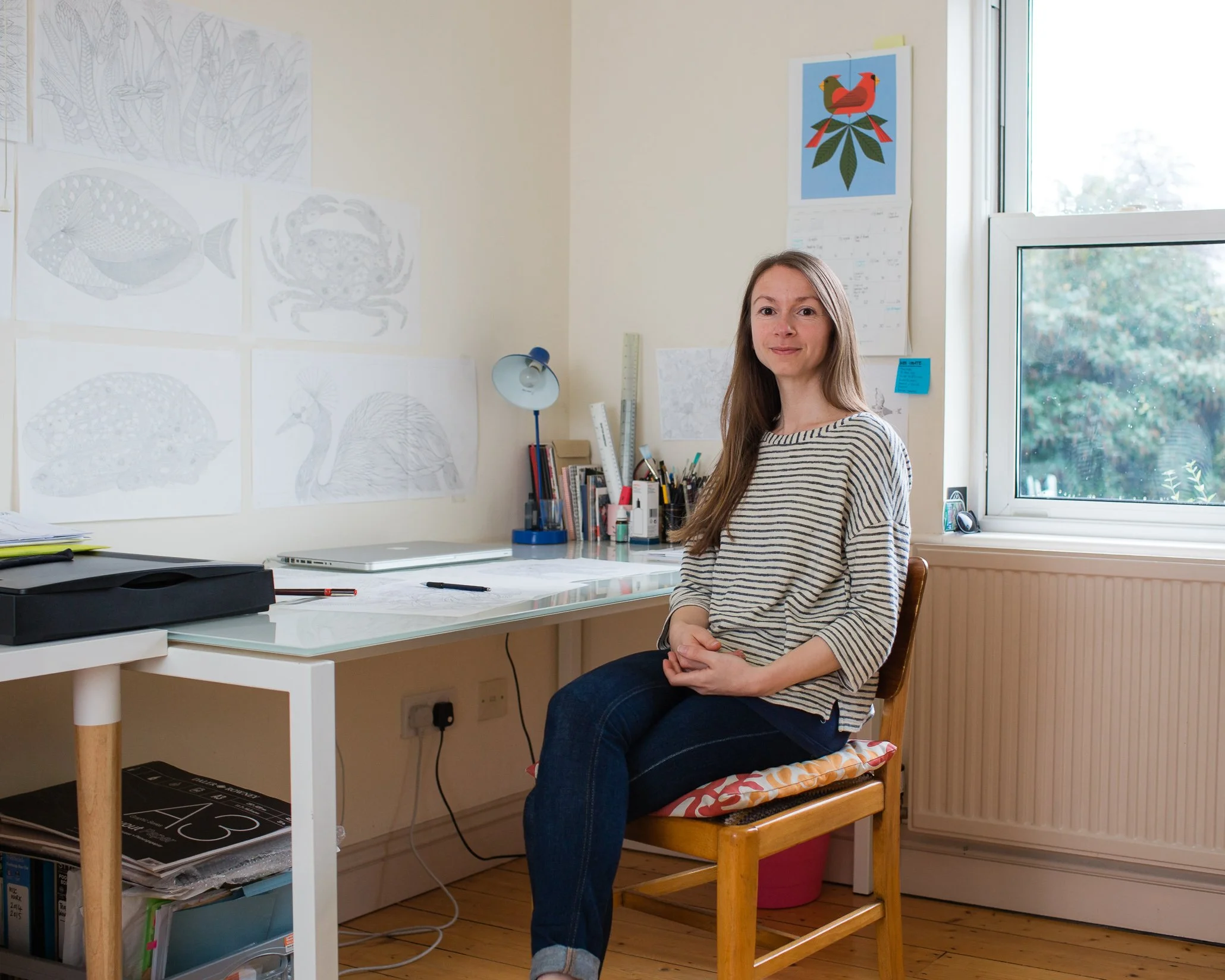 Woman sitting at a desk in an art studio, surrounded by sketches of animals and plants on the wall, with art supplies and books on the desk.