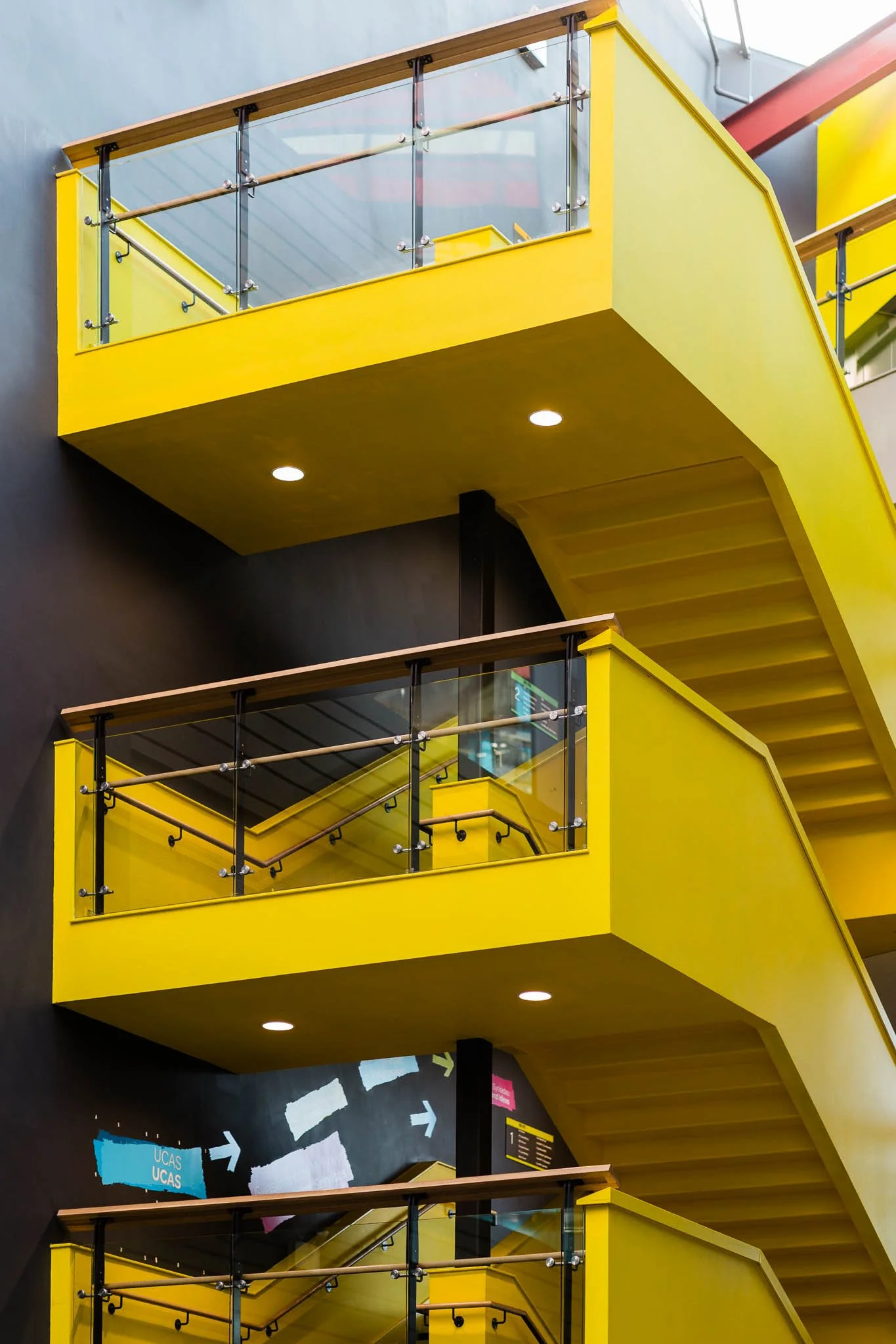 Multiple yellow staircases in an indoor area with glass railings and black railings, illuminated by recessed ceiling lights.