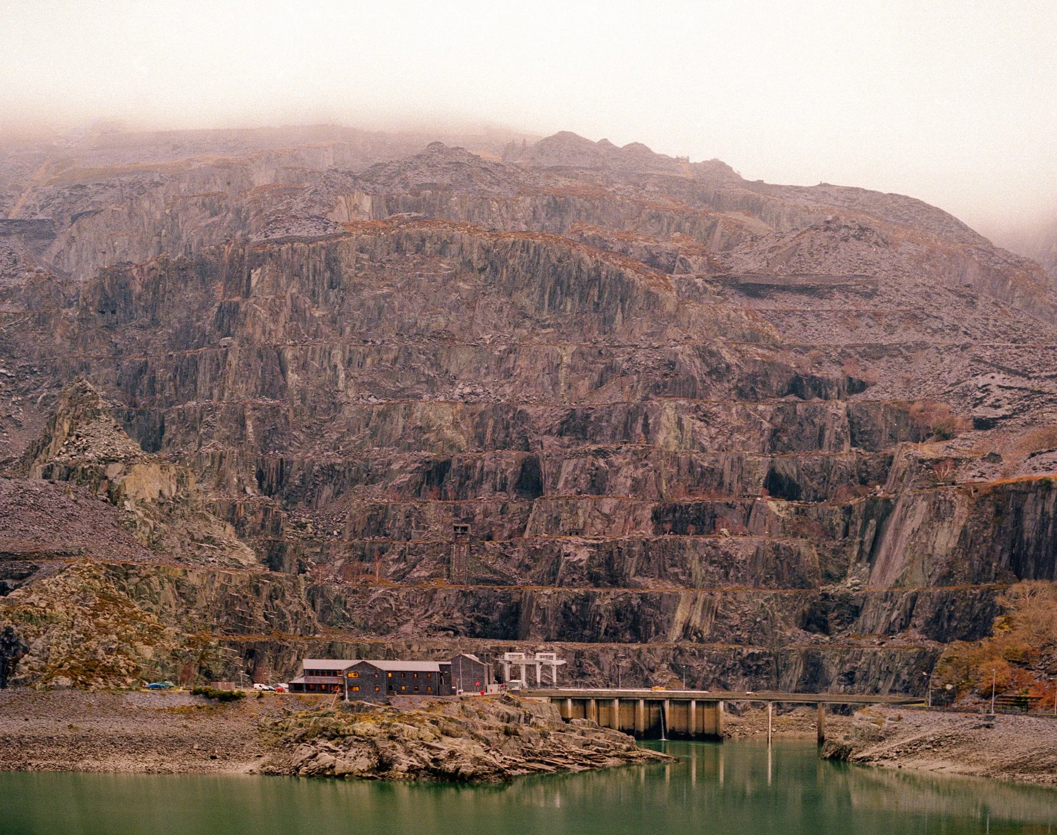 Hydroelectric dam in a canyon with water at the bottom, surrounded by rugged rocky mountains, and a building near the dam.