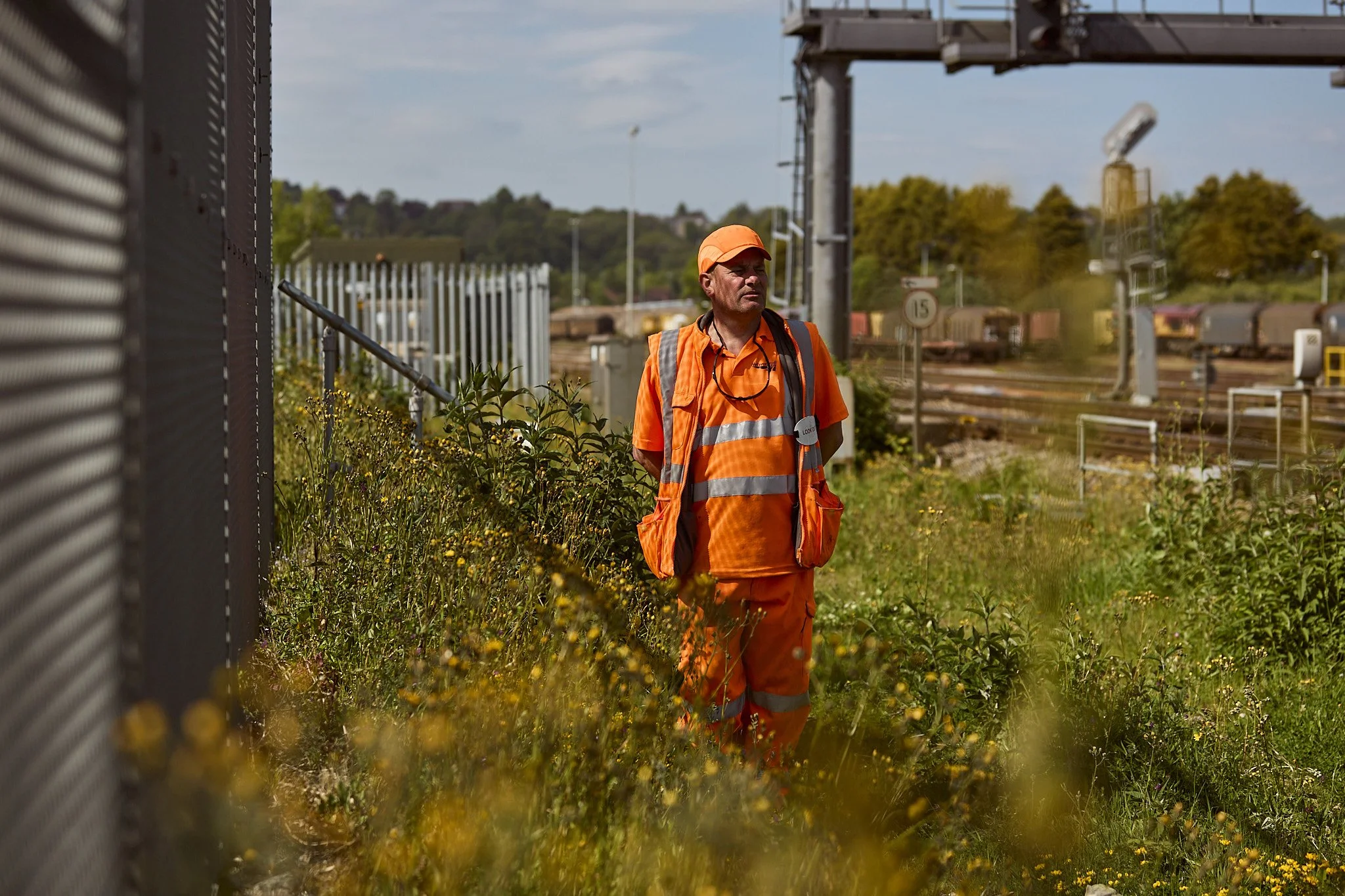 A man in orange construction work attire and a cap standing outdoors near railway tracks, surrounded by greenery and wildflowers, with trains and railway infrastructure in the background.