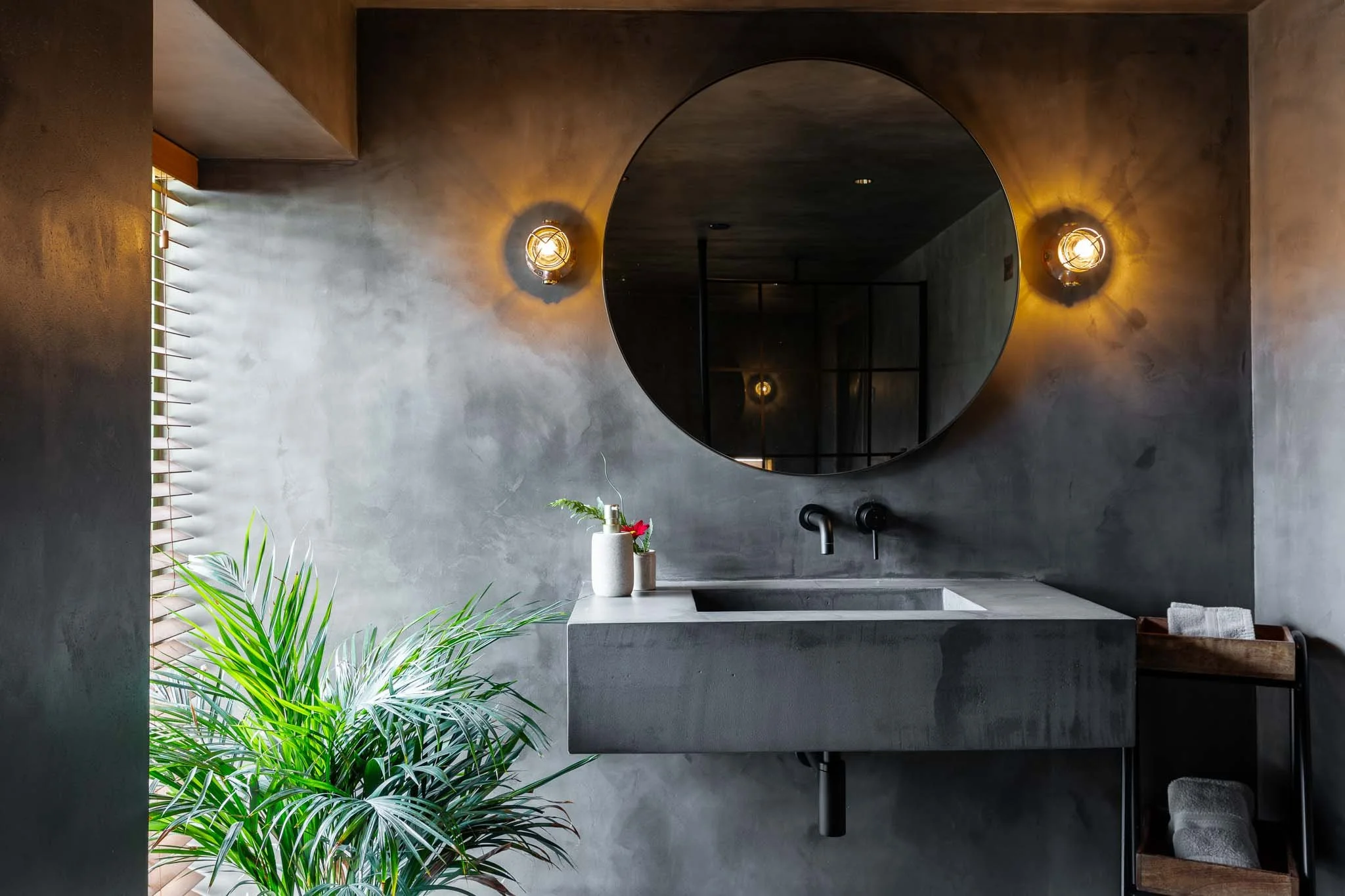 Modern bathroom with a concrete sink, round mirror, two wall-mounted lights, a streak of sunlight from a window with blinds, and green plants