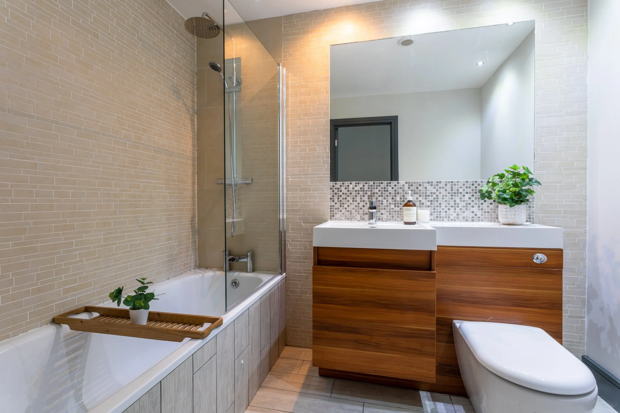 Modern bathroom with beige brick tiles, a bathtub with a wooden bath tray and potted plant, a glass-enclosed shower, a wooden vanity with a white sink, mirrored wall, and a decorative mosaic tile backsplash. There's also a white smart toilet and a green potted plant on the vanity.