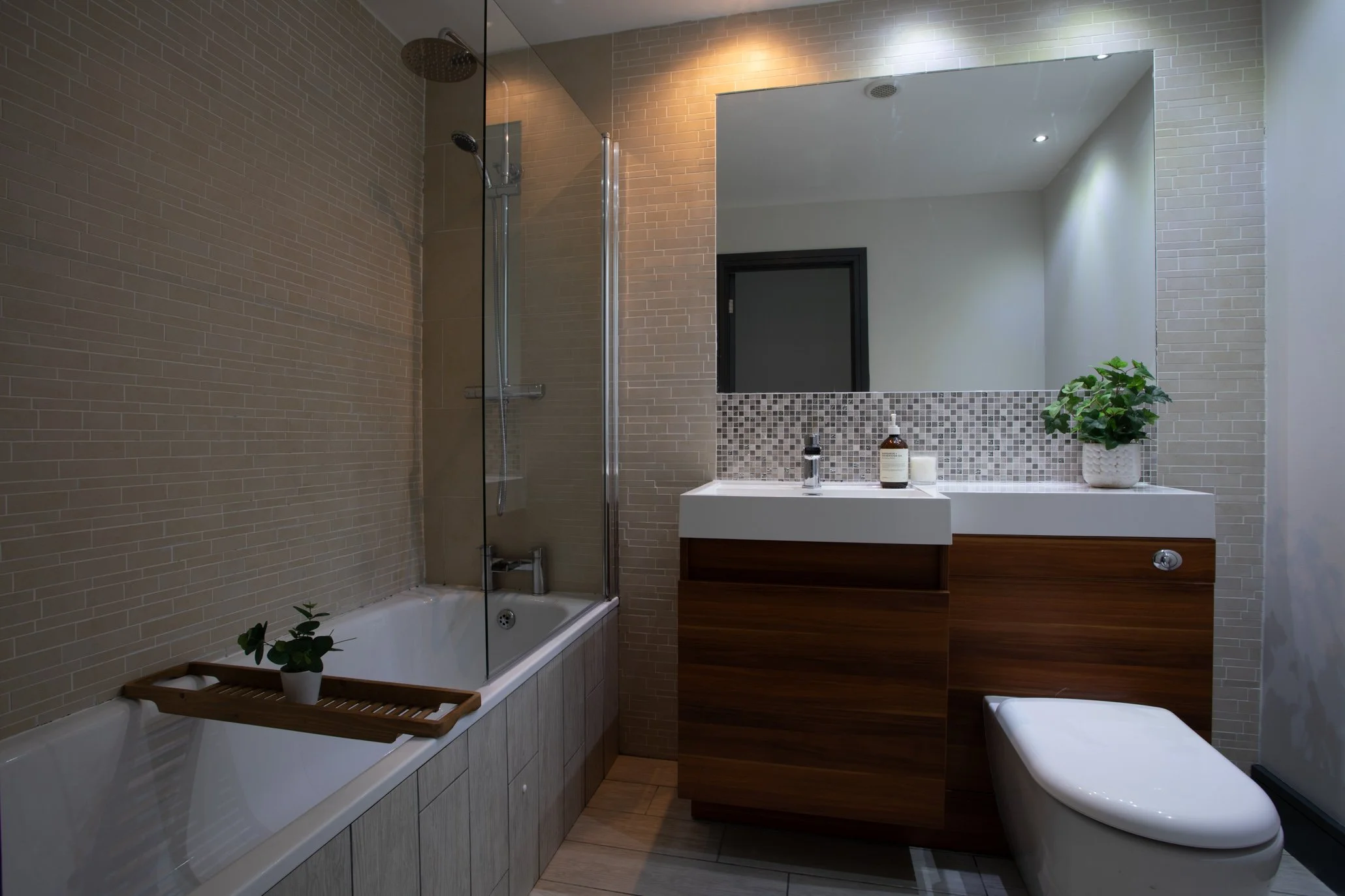 Modern bathroom with a bathtub, shower, sink with a wooden cabinet, and a toilet. The bathroom features beige tiles, a large mirror, and decorative mosaic tiles behind the sink. There are potted plants on the bathtub and near the sink.