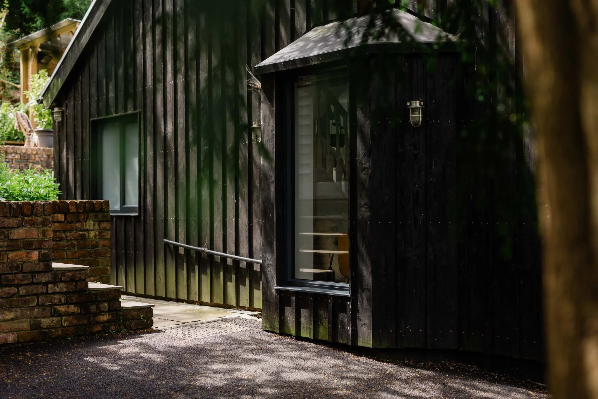A black wooden building with a small entry door and a large window, two outdoor wall lights, and a brick pathway with steps leading up to it, surrounded by greenery.