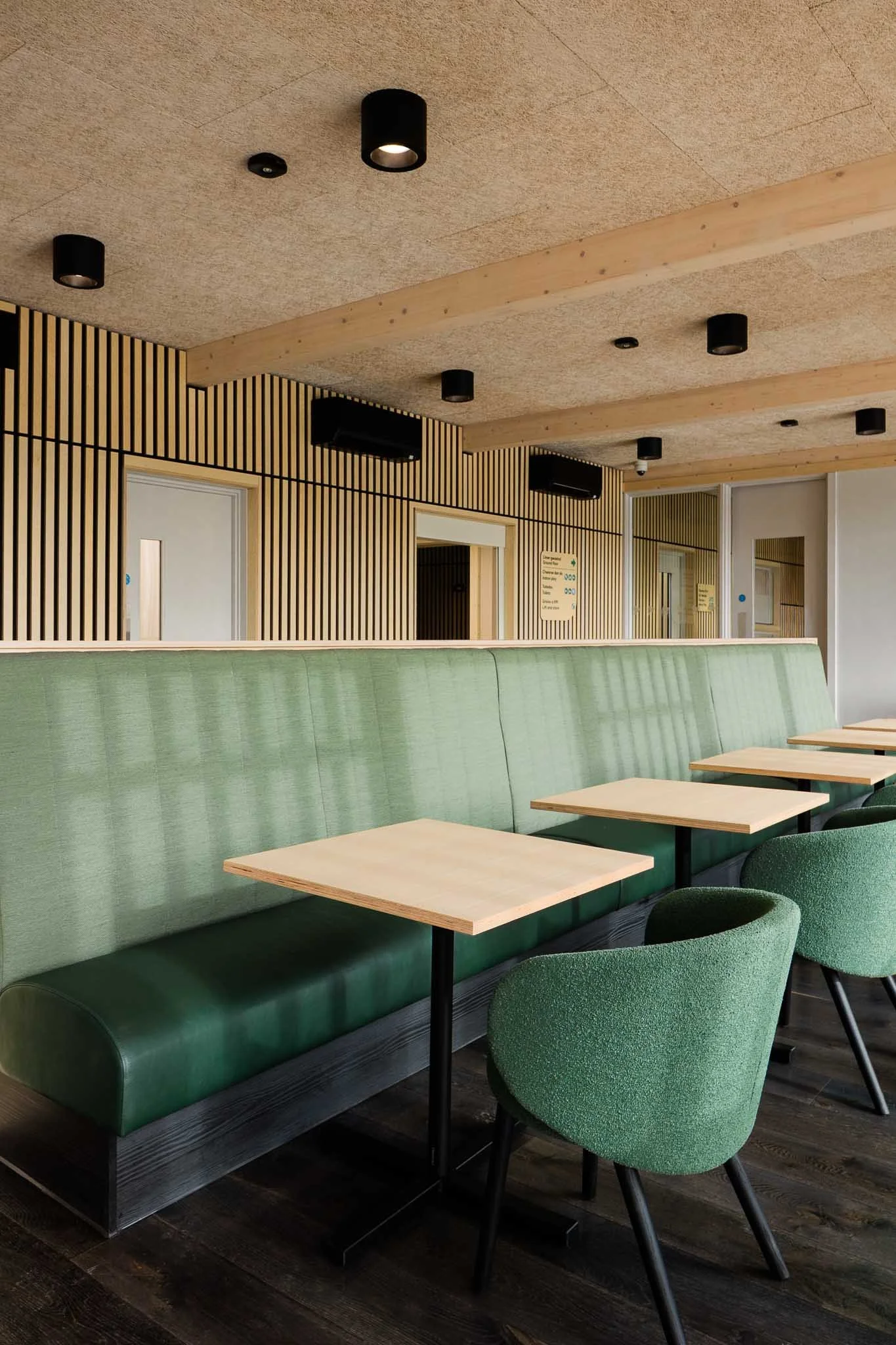 Interior of a modern cafe or lounge with green upholstered seating, light wooden tables, and green chairs, with wooden slat wall accents and black ceiling lights.