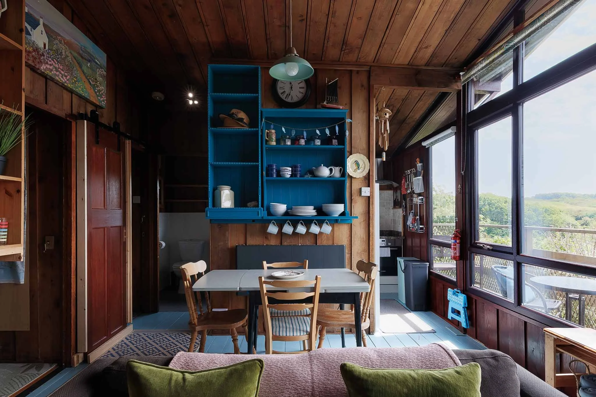 Indoor dining area with wooden walls and ceiling, large windows with views of greenery, a blue open shelf with dishes, a small round dining table with chairs, and various decorative items.