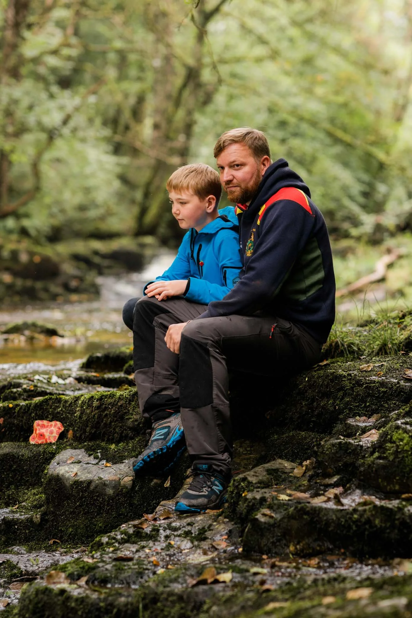 An adult male and a young boy sitting on a mossy rock by a stream in a forest, both dressed in outdoor hiking clothes.